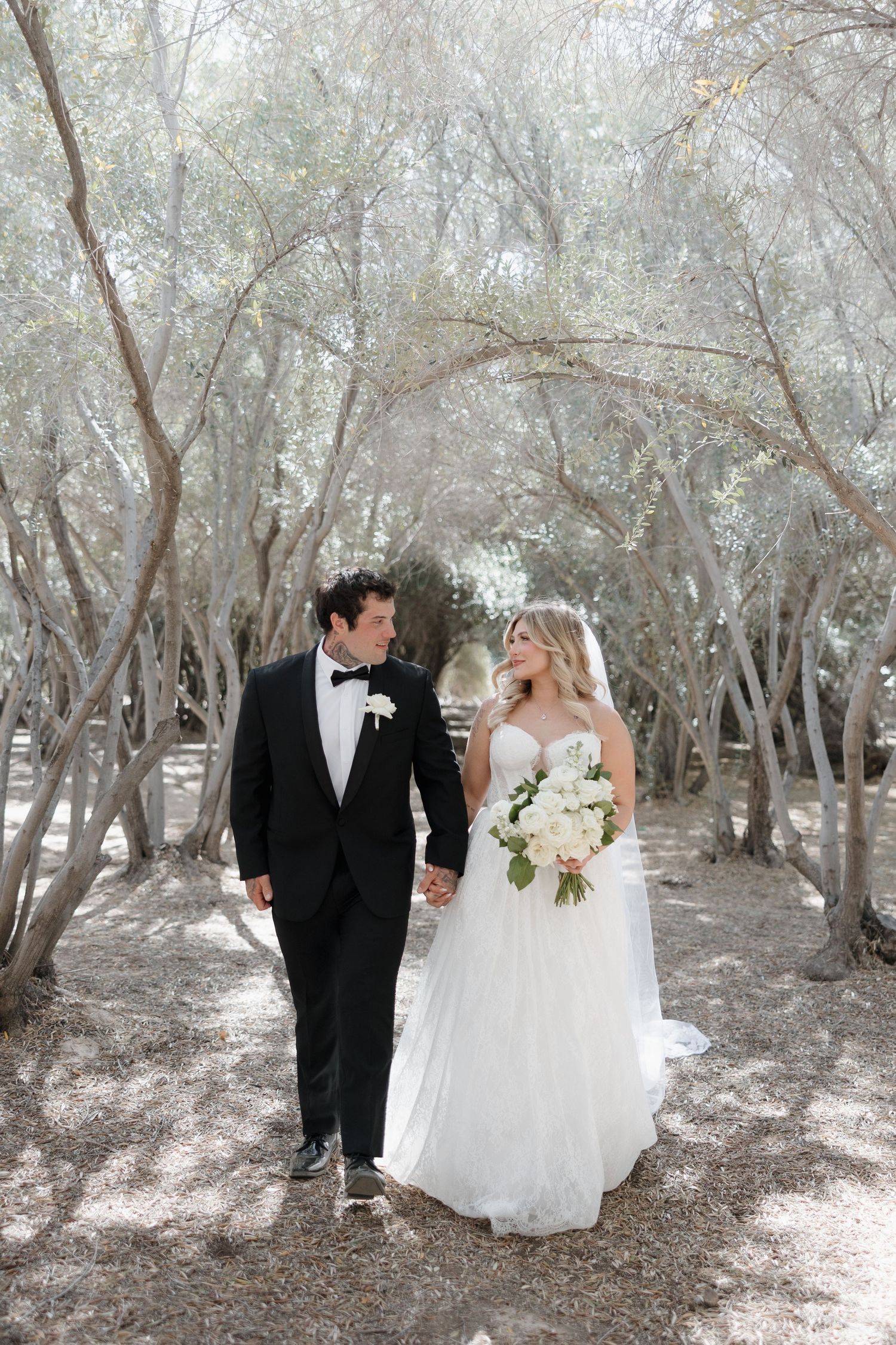 Bride and groom walk hand in hand through arched silver trees, bride holds white bouquet.