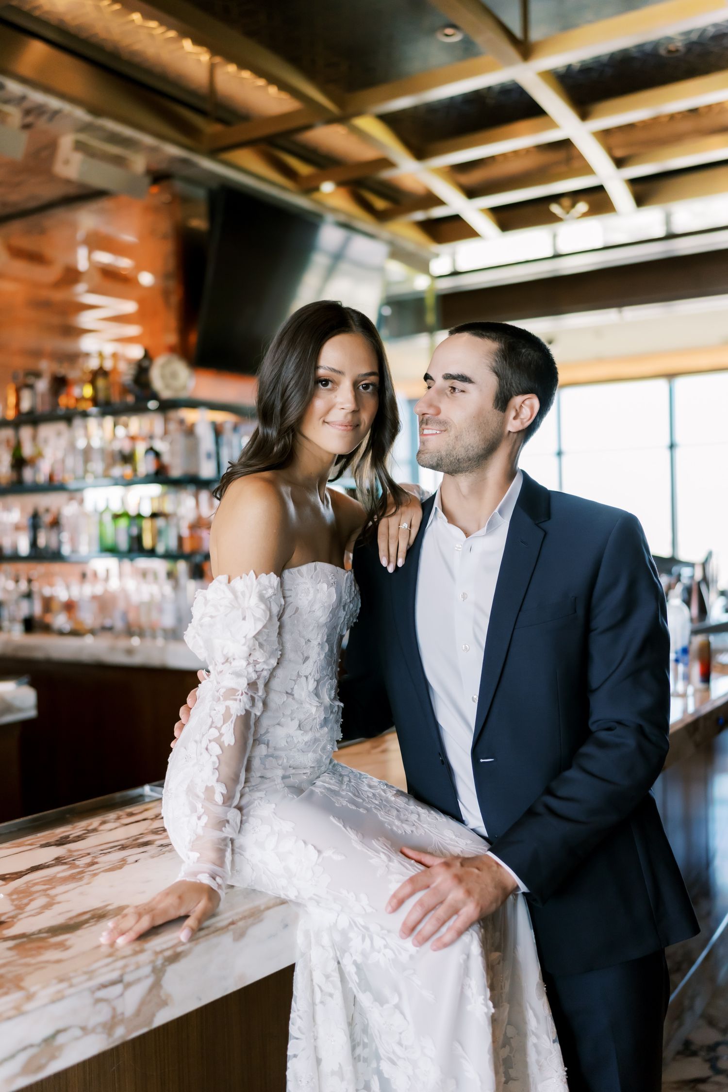 Bride in lace off-shoulder gown and groom in navy suit pose together at an elegant marble bar.