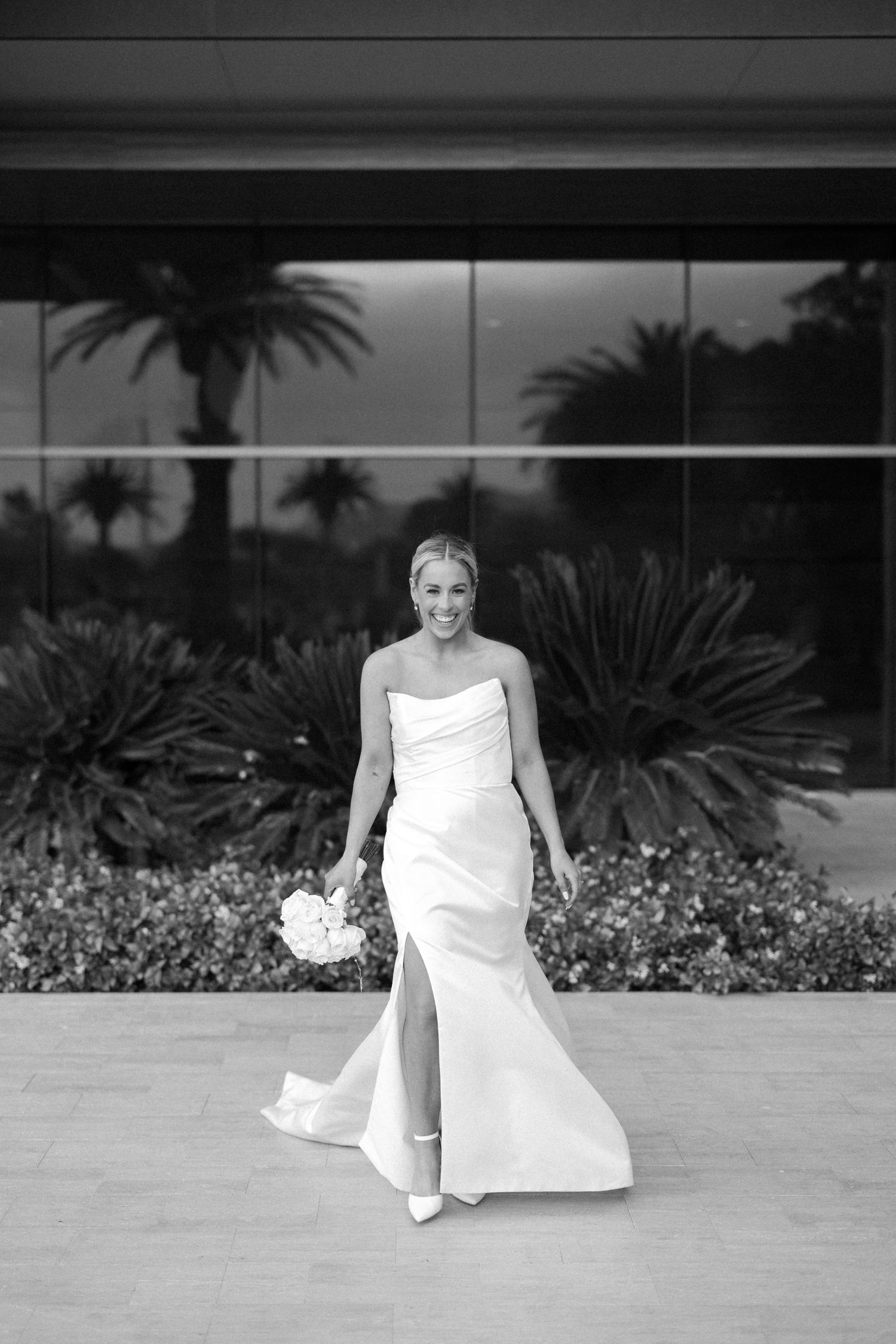 Smiling bride in strapless white gown with slit holds bouquet outdoors near lush greenery and modern glass building.