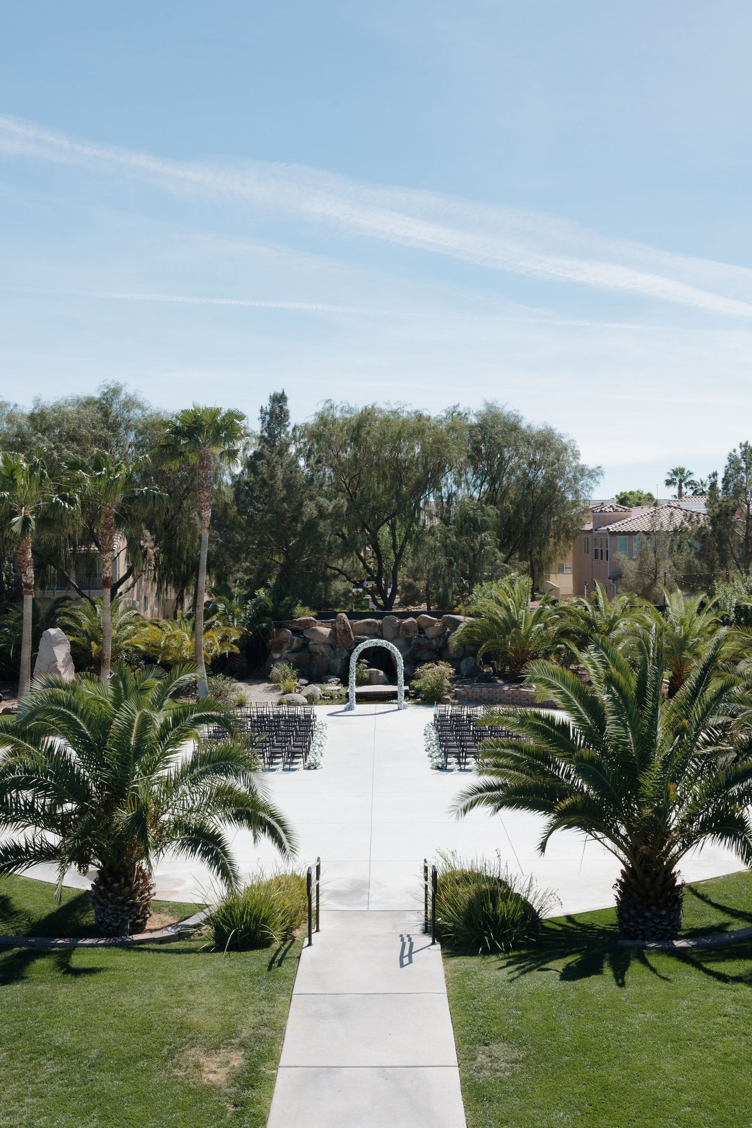 Outdoor wedding ceremony setup with rows of chairs, white arch, palm trees, and lush greenery under a clear blue sky.