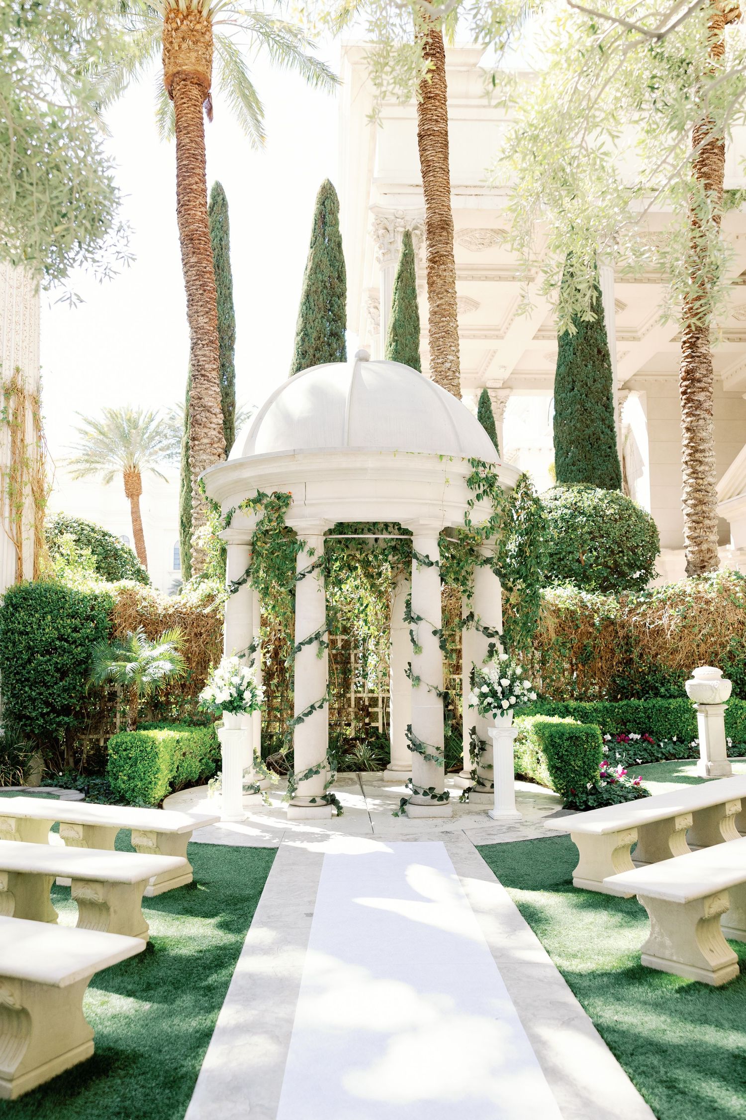 Elegant outdoor wedding ceremony venue with white gazebo, ivy, manicured hedges, stone benches, and tall cypress trees.