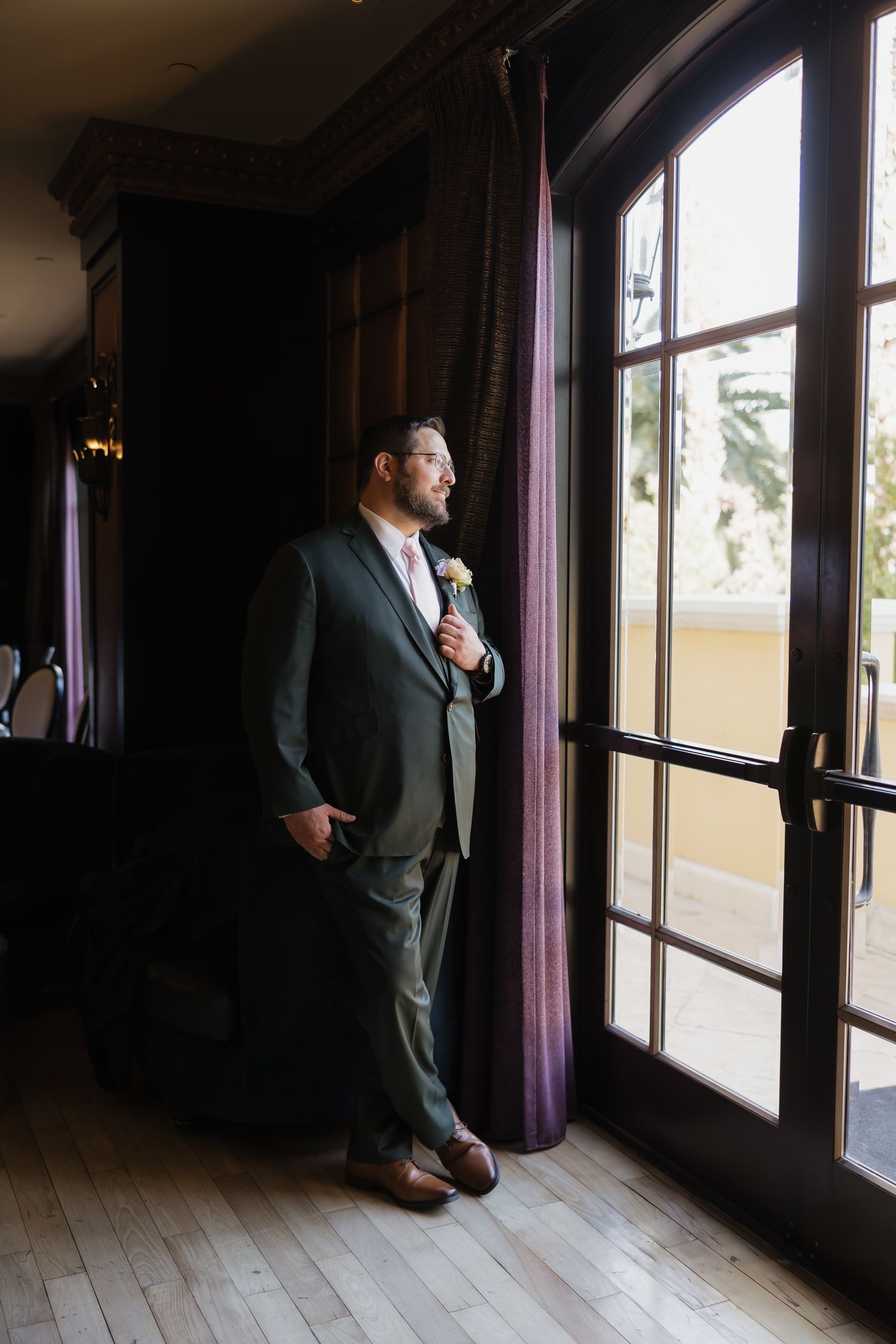 A dapper groom in a gray suit with boutonniere stands by French doors with purple curtains on his wedding day.