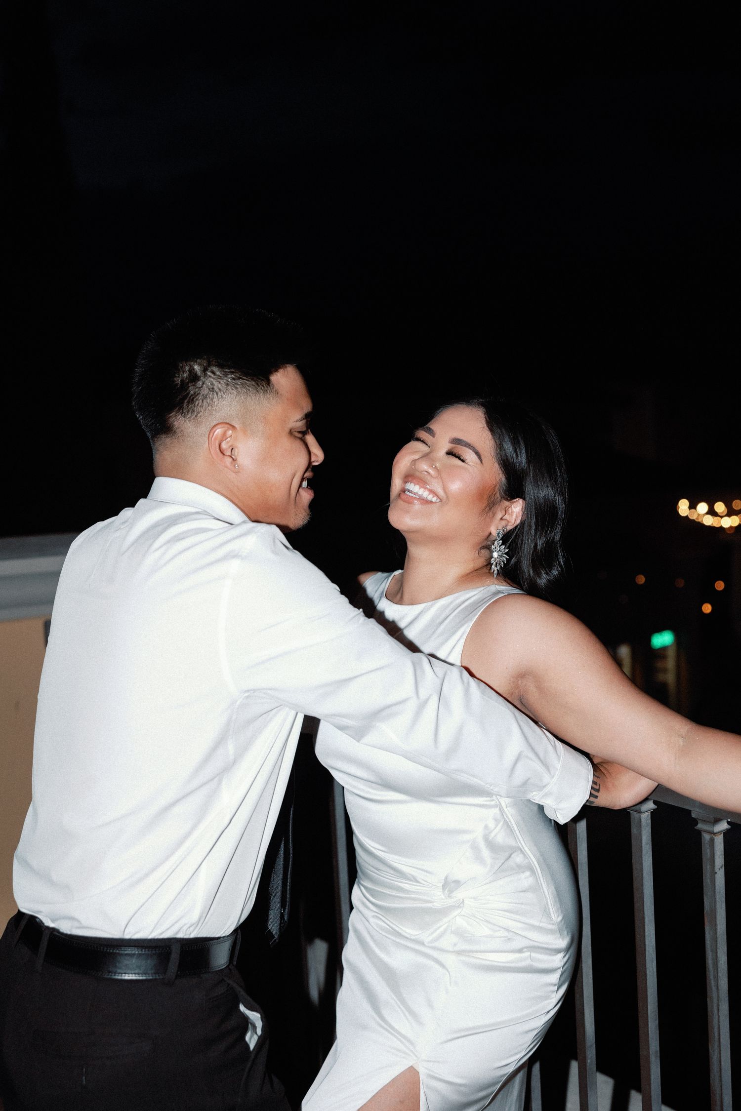 Couple in white outfits smiling and embracing on a balcony at night.