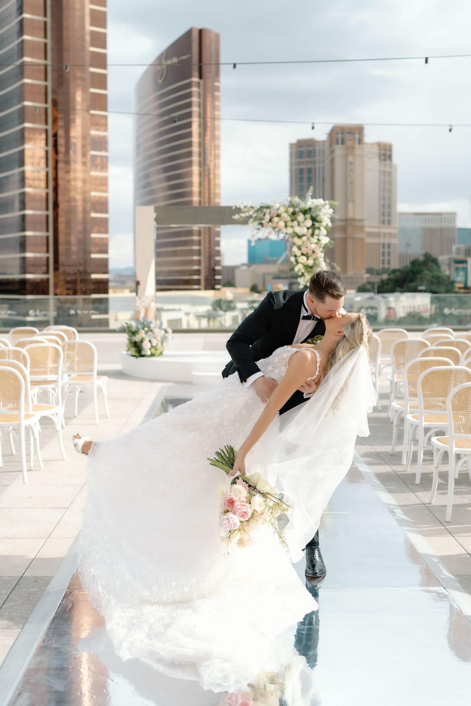 Bride and groom share a romantic dip kiss on a rooftop wedding venue with city skyline backdrop and floral arch.