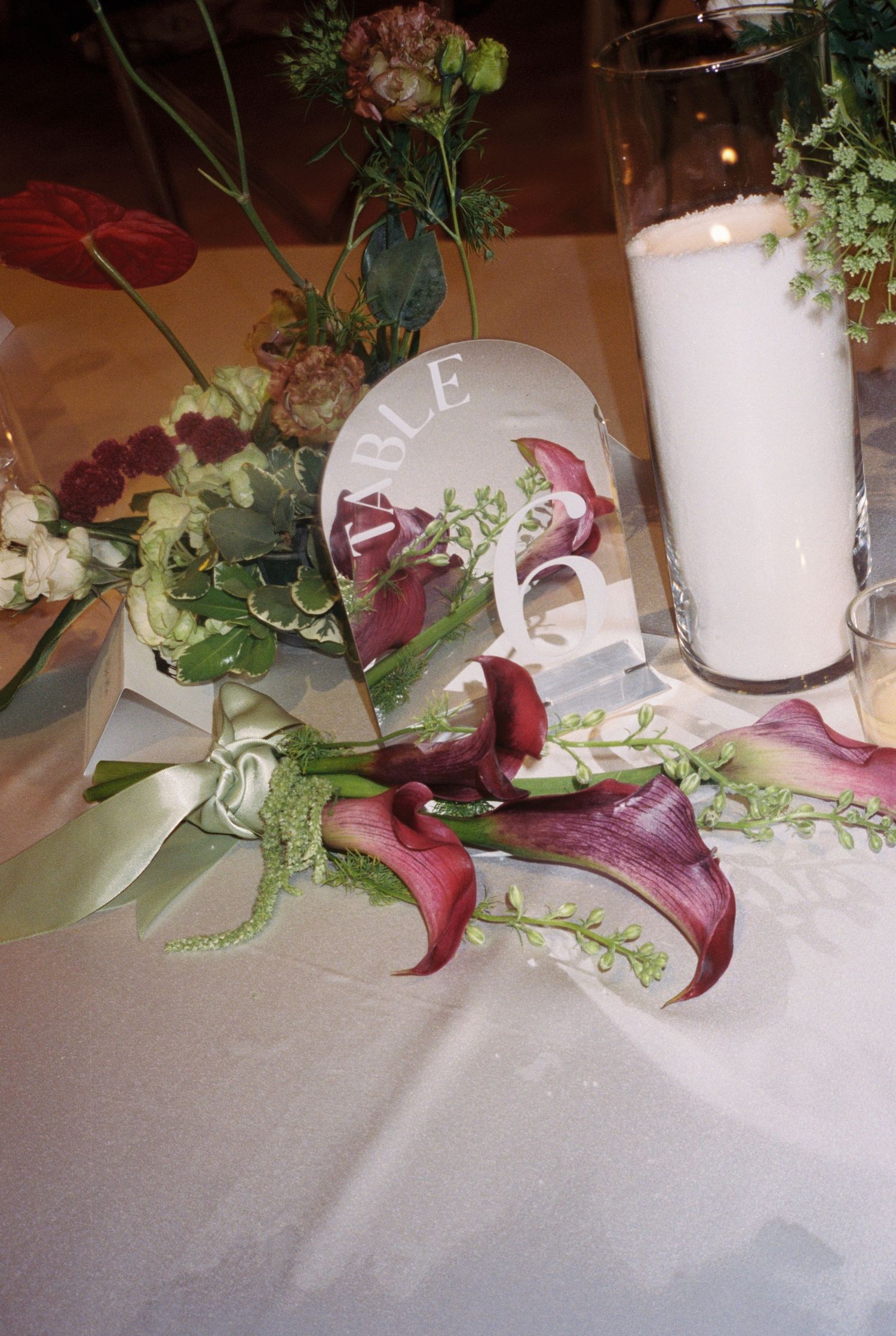 Elegant wedding table centerpiece with burgundy calla lilies, mixed florals, greenery, a candle, and a mirrored table number sign.