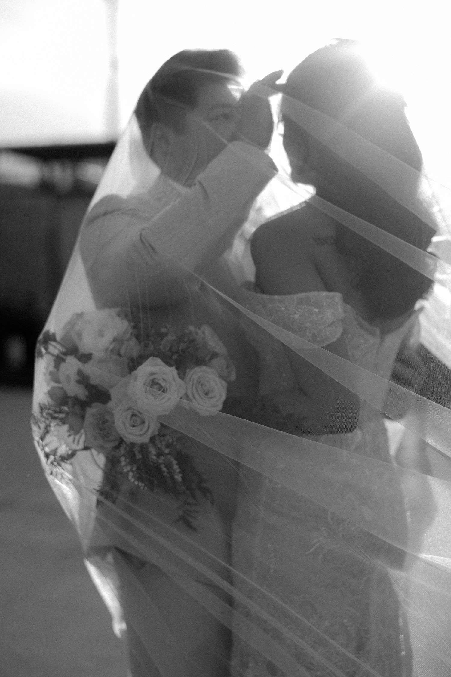 Black and white photo of bride and groom sharing romantic moment under flowing veil, bride holding bouquet of roses.