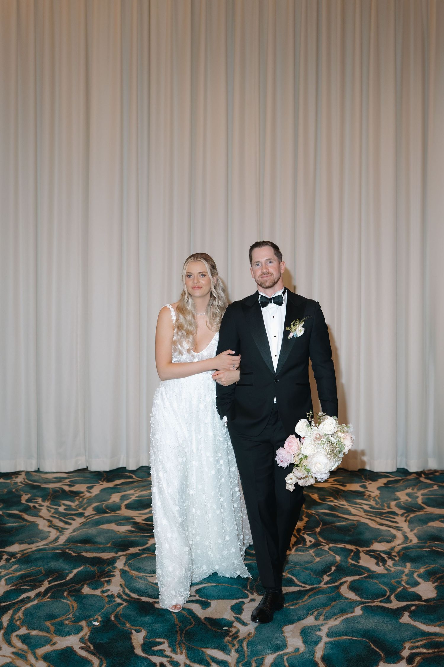 Bride and groom posing together in elegant wedding attire, she holds a pink and white bouquet.