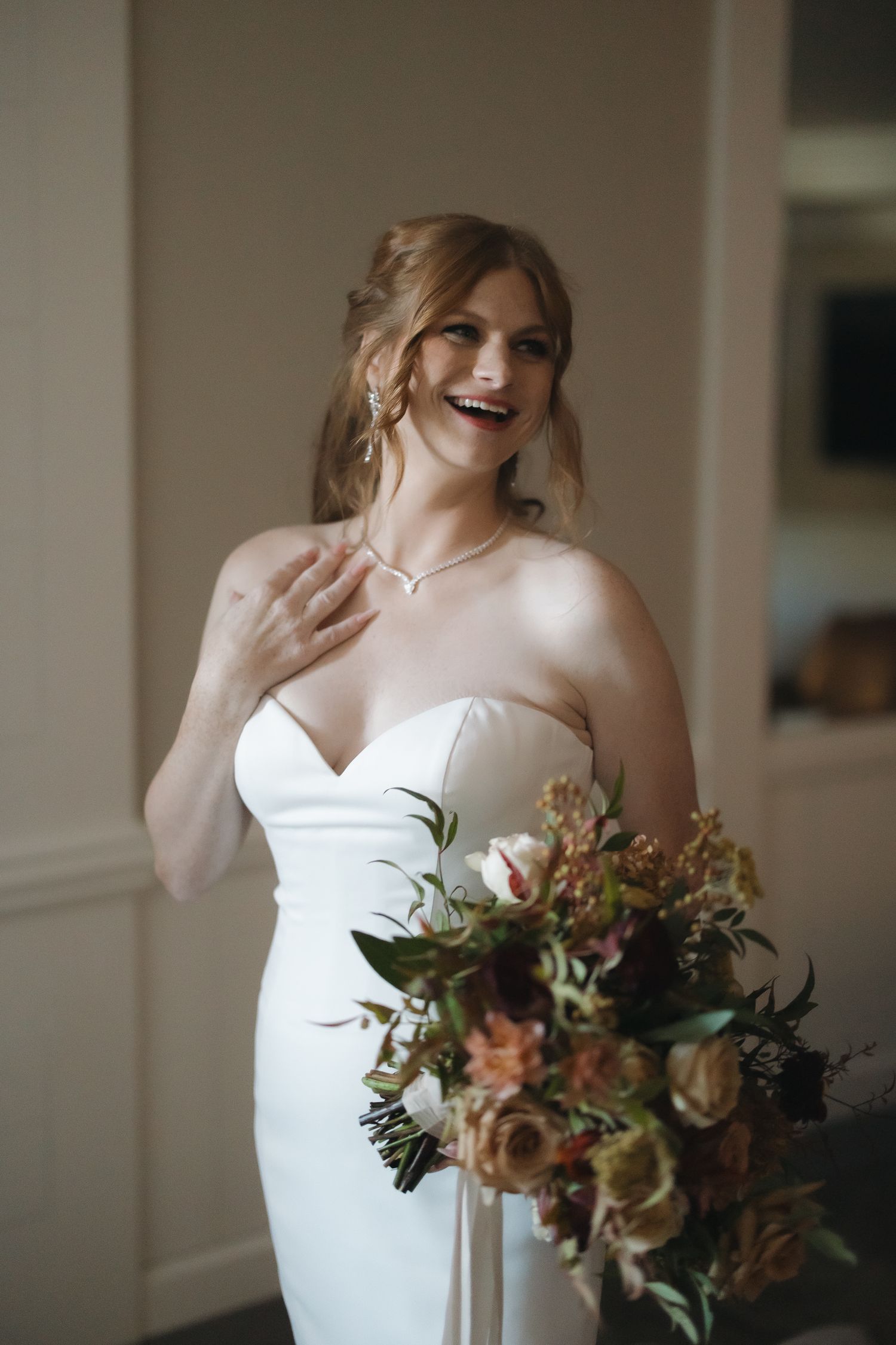 Radiant bride in strapless white gown holding autumn bouquet, laughing joyfully while touching her necklace.