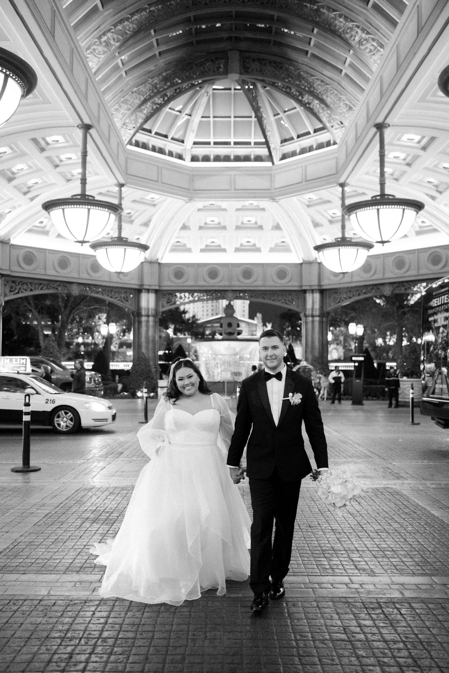 Bride and groom walk hand-in-hand under an ornate glass atrium in a black and white Las Vegas wedding photo.