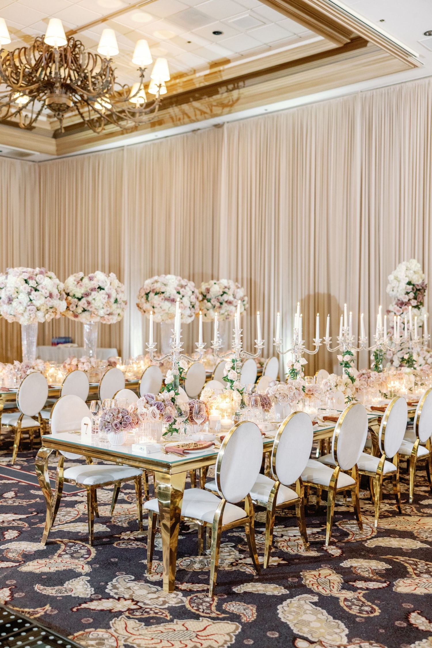Luxurious ballroom wedding reception with gold-trimmed white chairs and tall floral centerpieces.