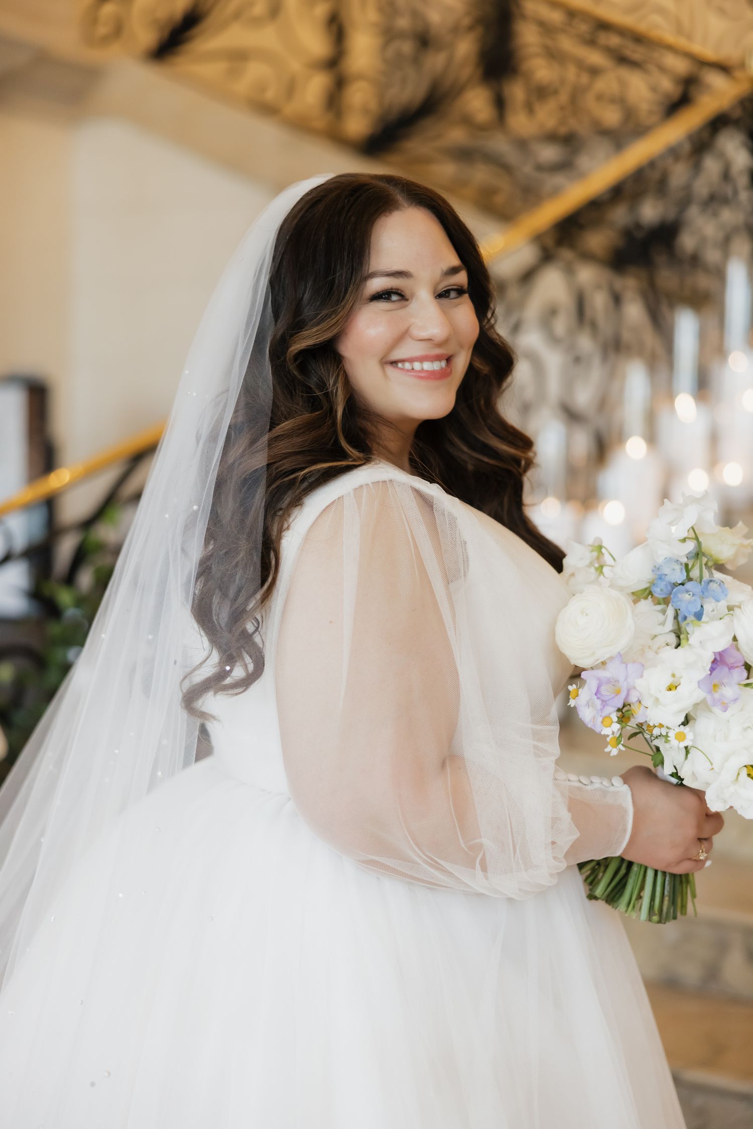 Smiling bride in white tulle gown and veil holds blue and white bouquet indoors.