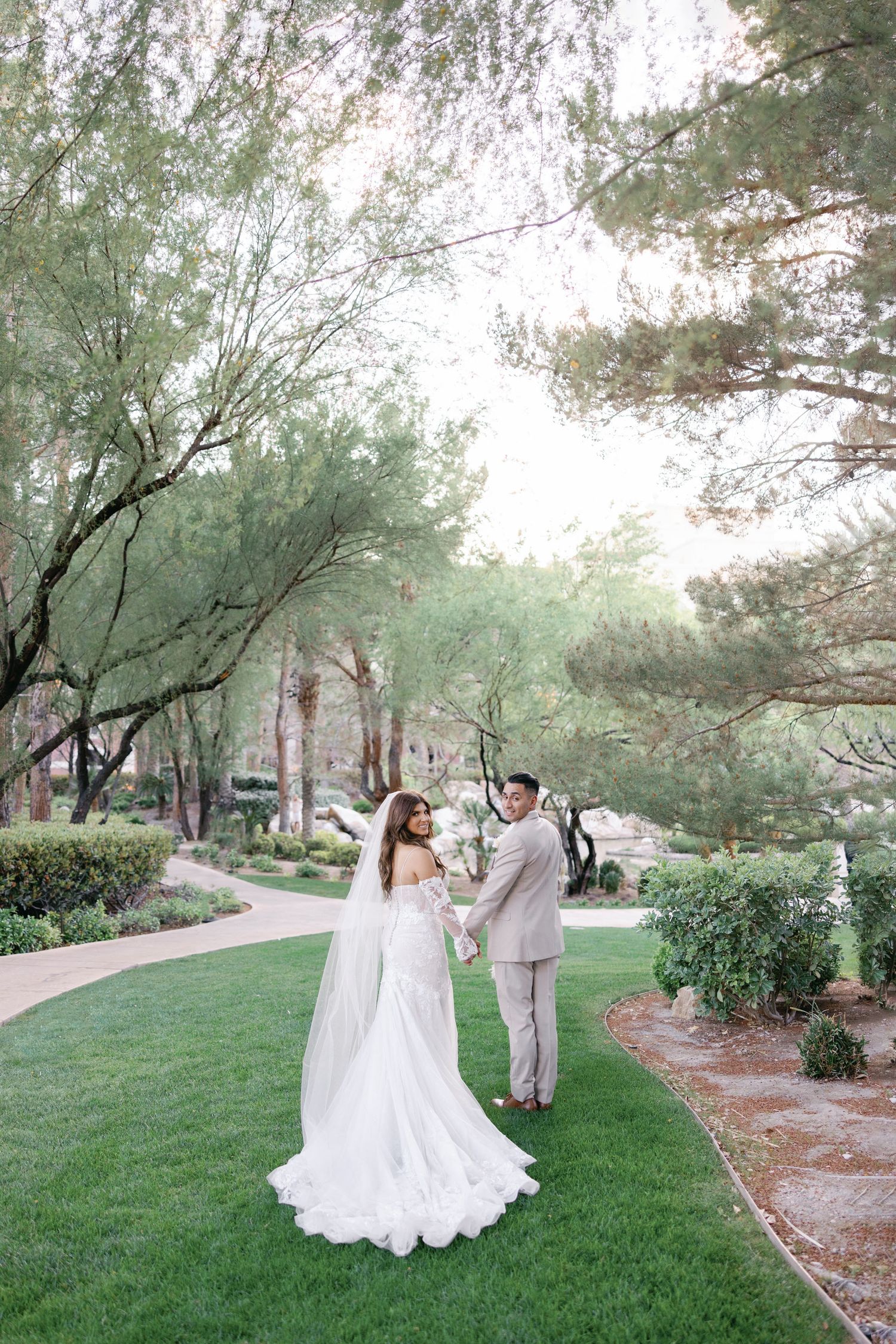 Bride and groom hold hands on a green garden path surrounded by tall elegant trees.