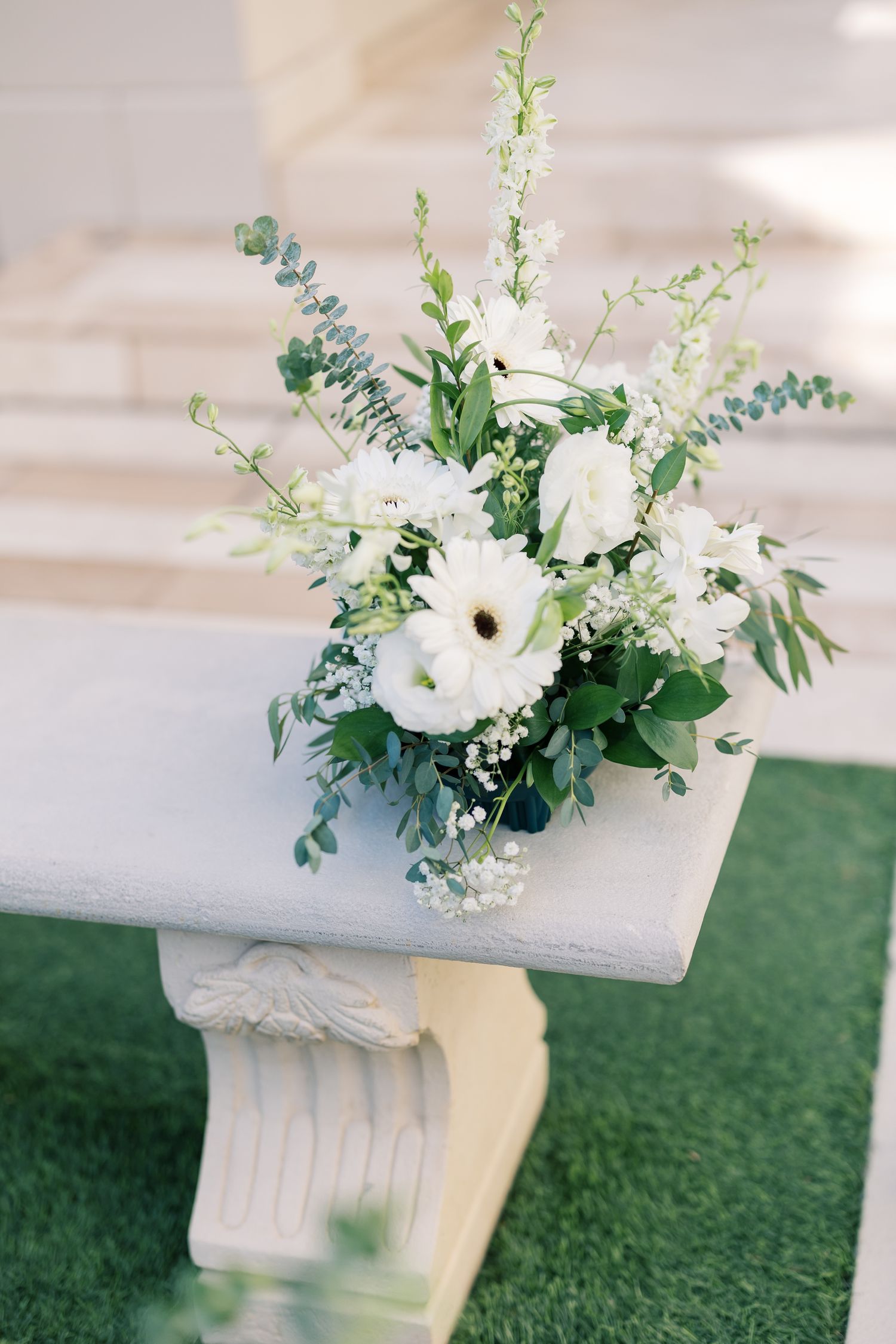 White floral arrangement with greenery on a stone bench at an outdoor wedding ceremony.