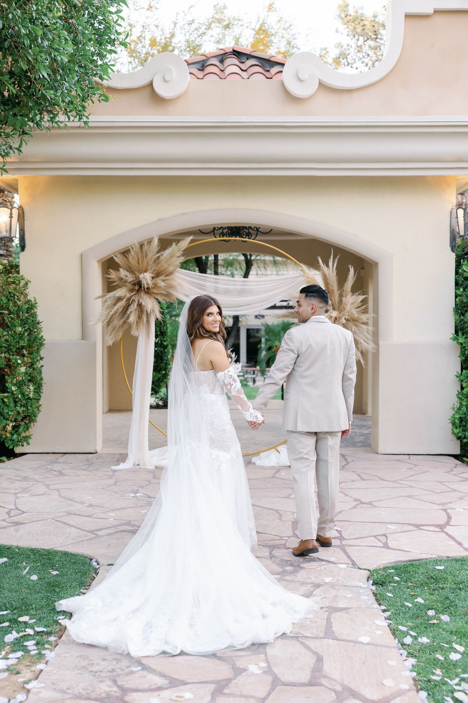 Bride and groom holding hands walking through floral arch with pampas grass at outdoor wedding venue.