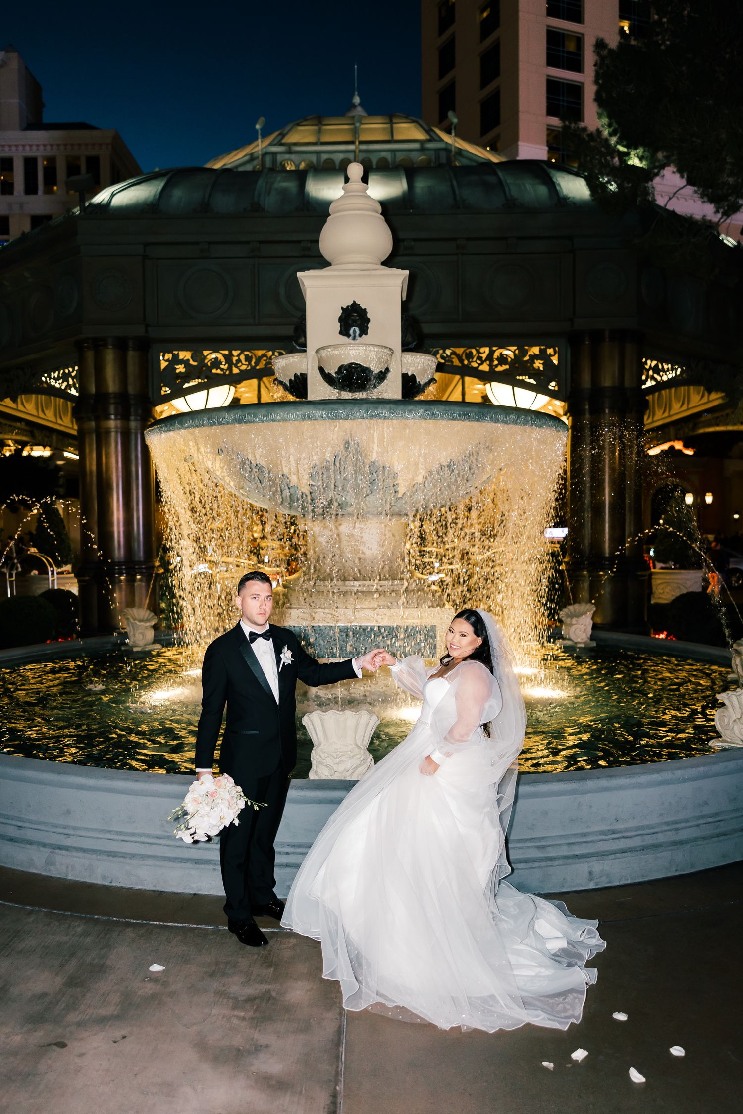 Bride and groom holding hands in front of a glowing ornate fountain at night in Las Vegas.