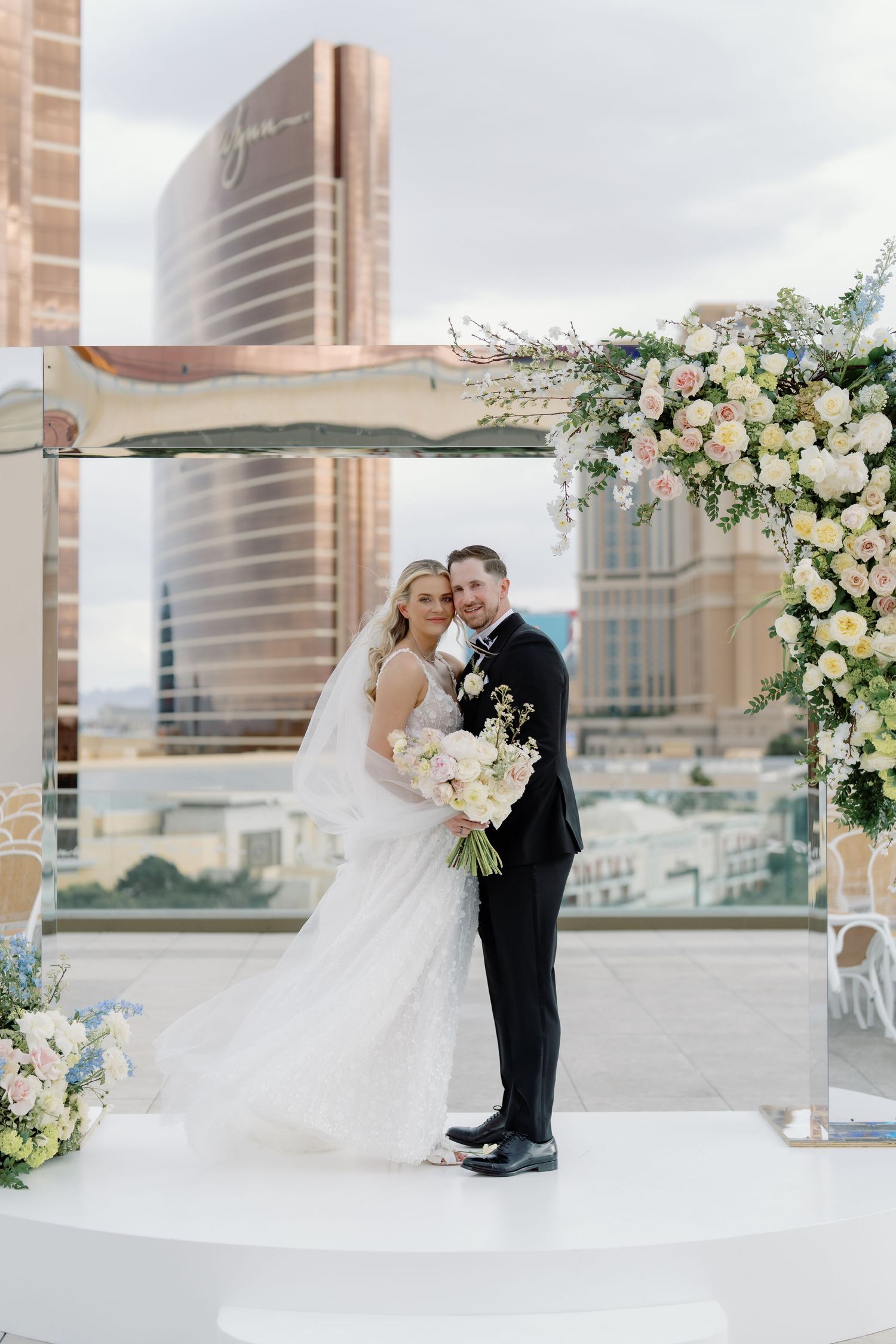 Bride and groom pose under floral arch on rooftop venue with Las Vegas skyline backdrop.