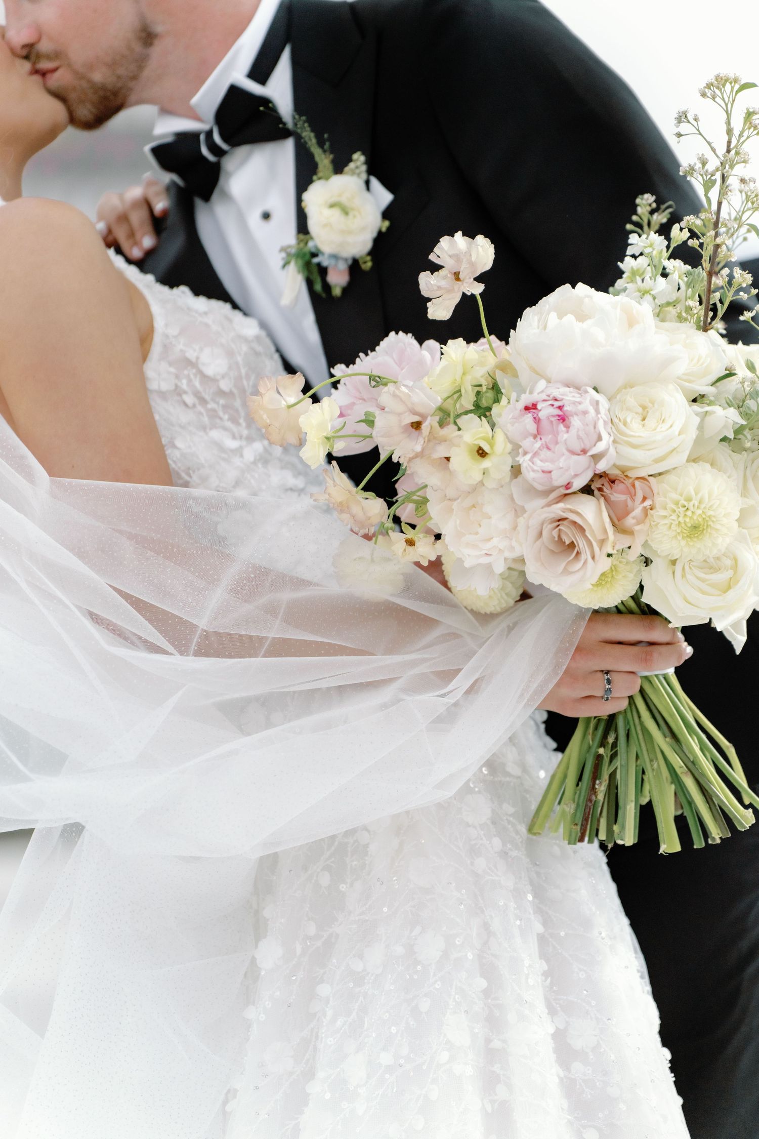 A bride and groom share a kiss, with the bride holding a soft pastel bouquet of peonies and roses, her veil flowing gracefully.