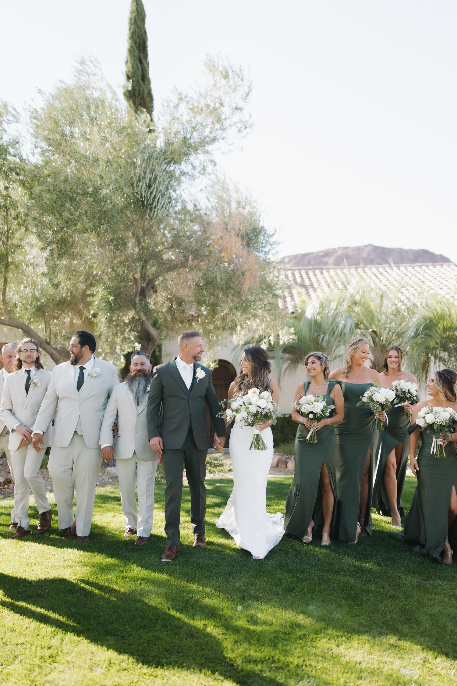 Wedding party walks outdoors, bride in white gown, bridesmaids in green dresses, groomsmen in gray suits, lush garden backdrop.