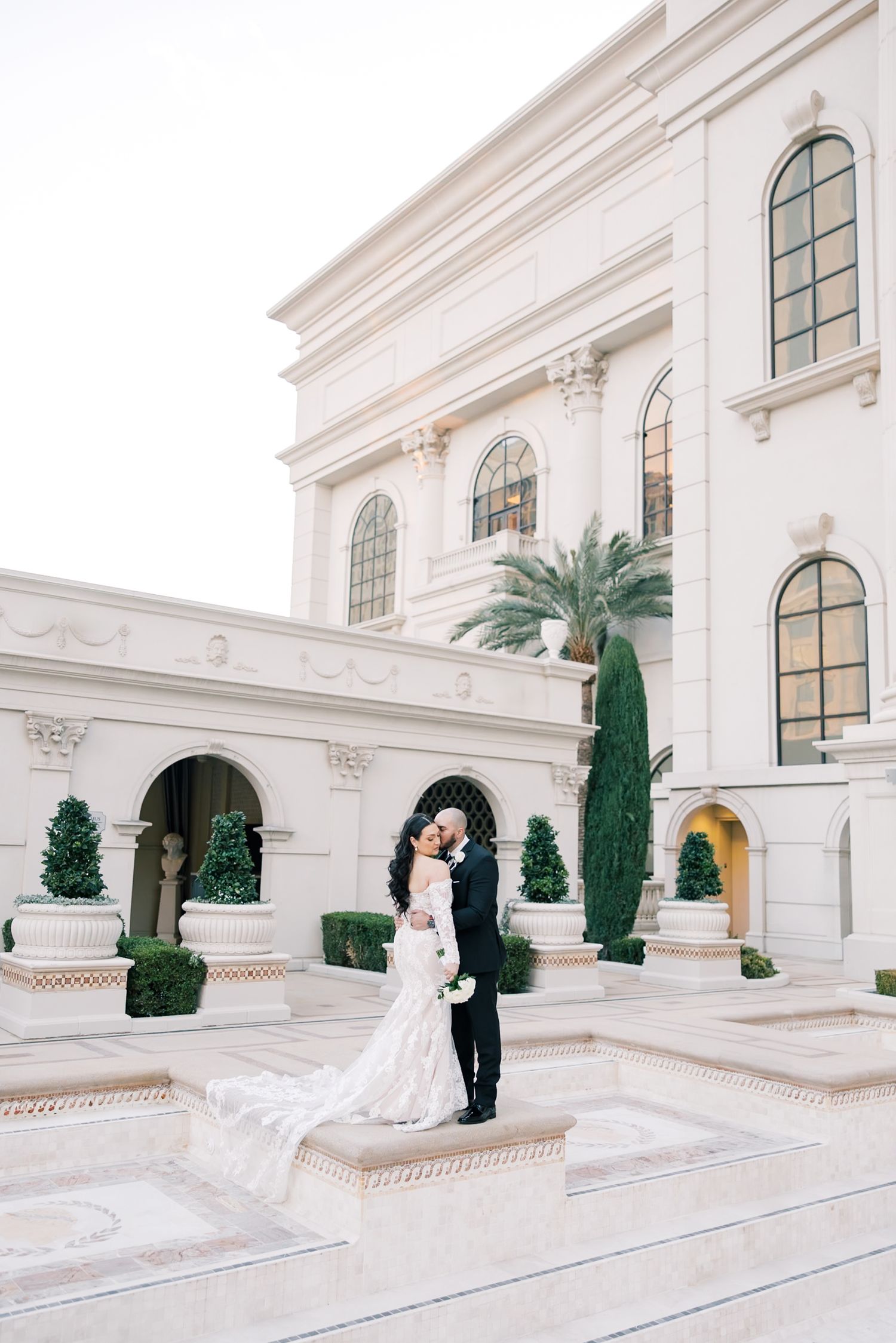 Bride and groom embracing in front of grand white neoclassical building with manicured hedges and palms.