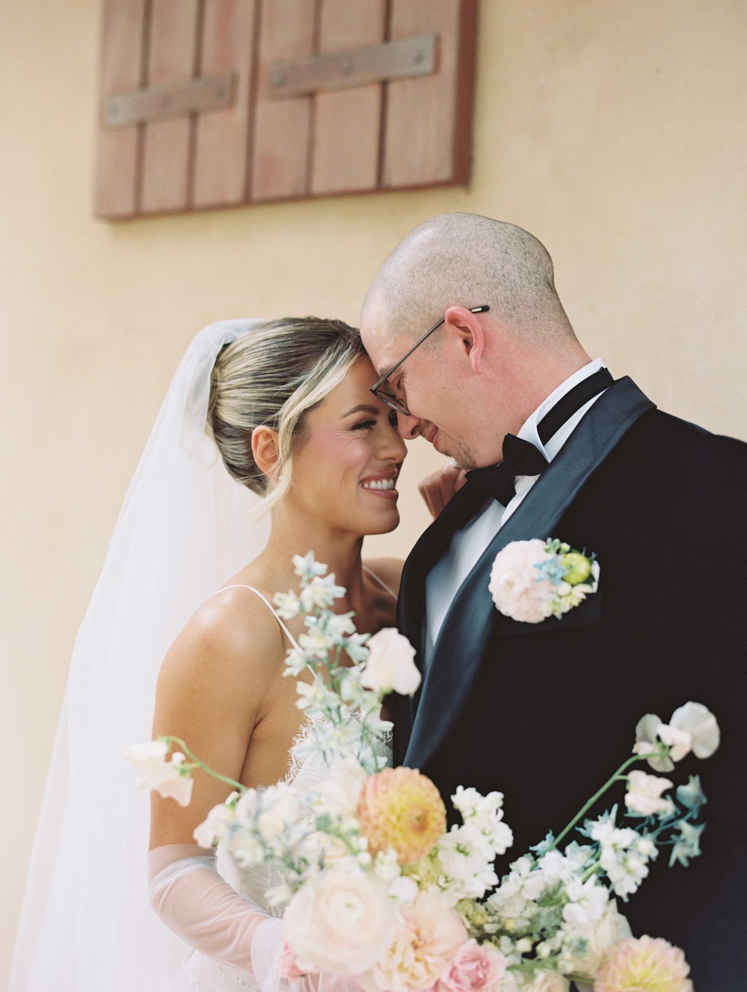 A bride and groom share a romantic moment, smiling forehead-to-forehead, with a pastel floral bouquet and wedding veil.