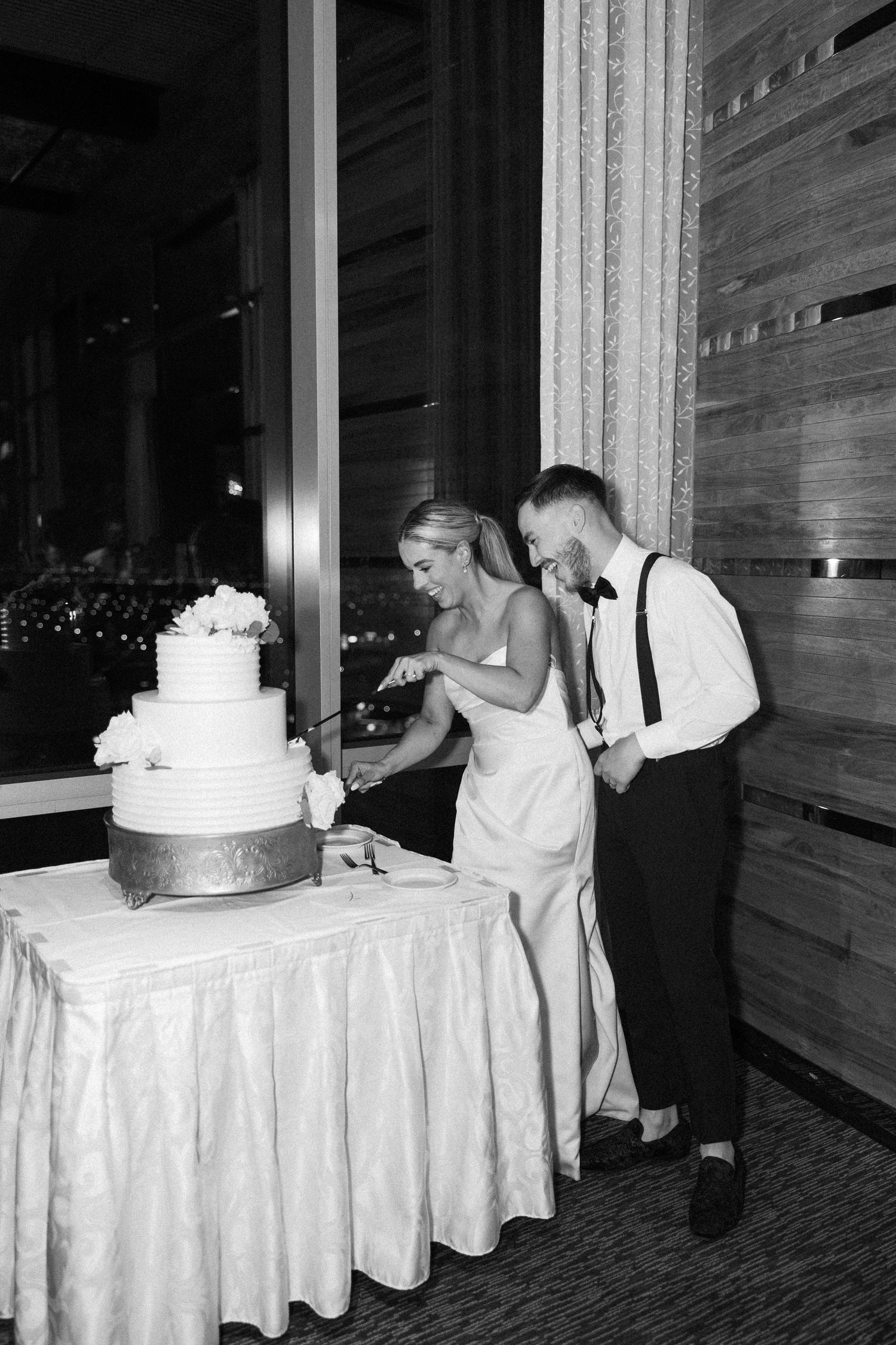Bride and groom cutting tiered white wedding cake at evening reception in black and white photo.