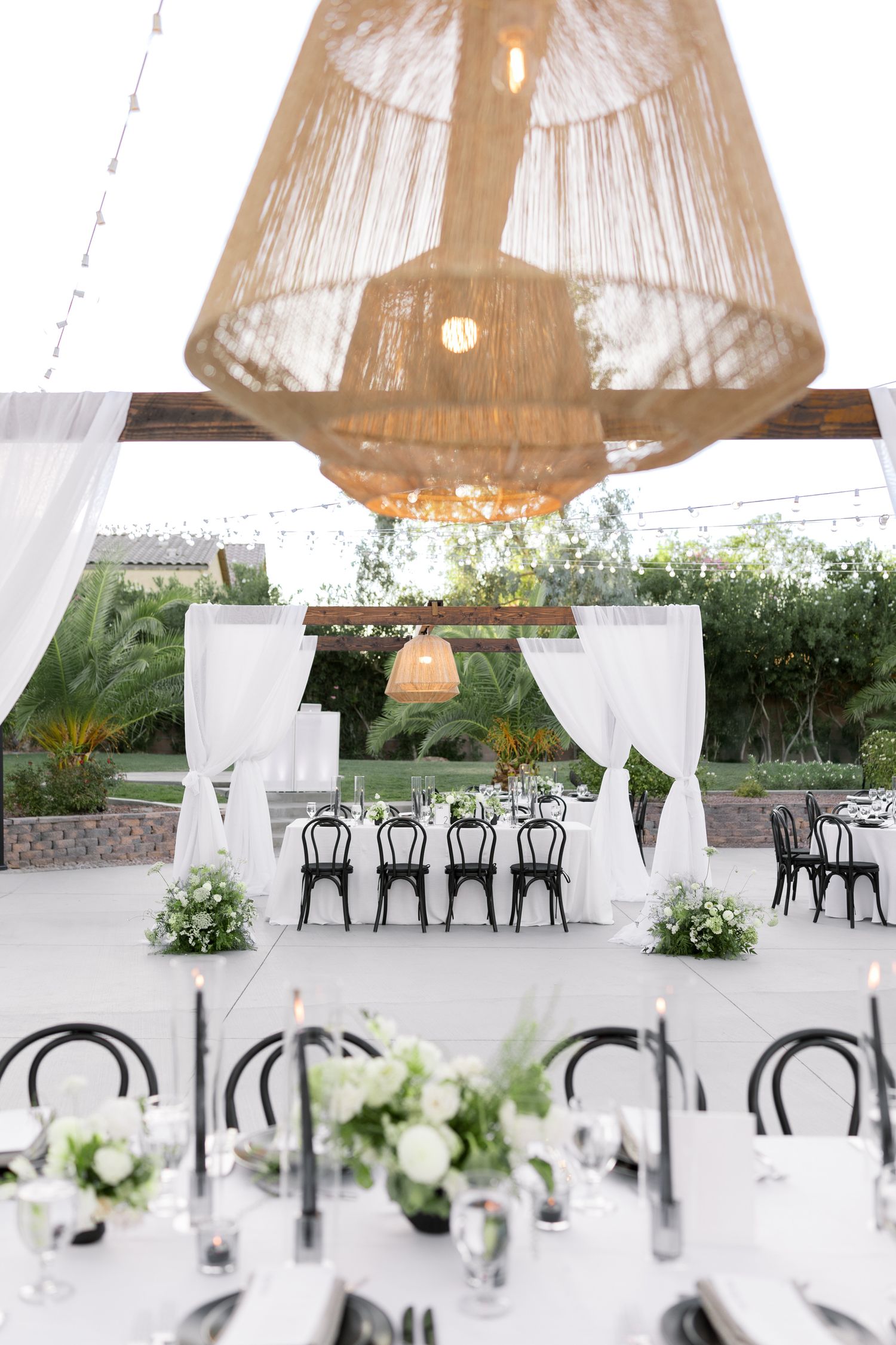 Elegant white wedding reception setup with wicker pendant lights and black chairs under a draped tent canopy.