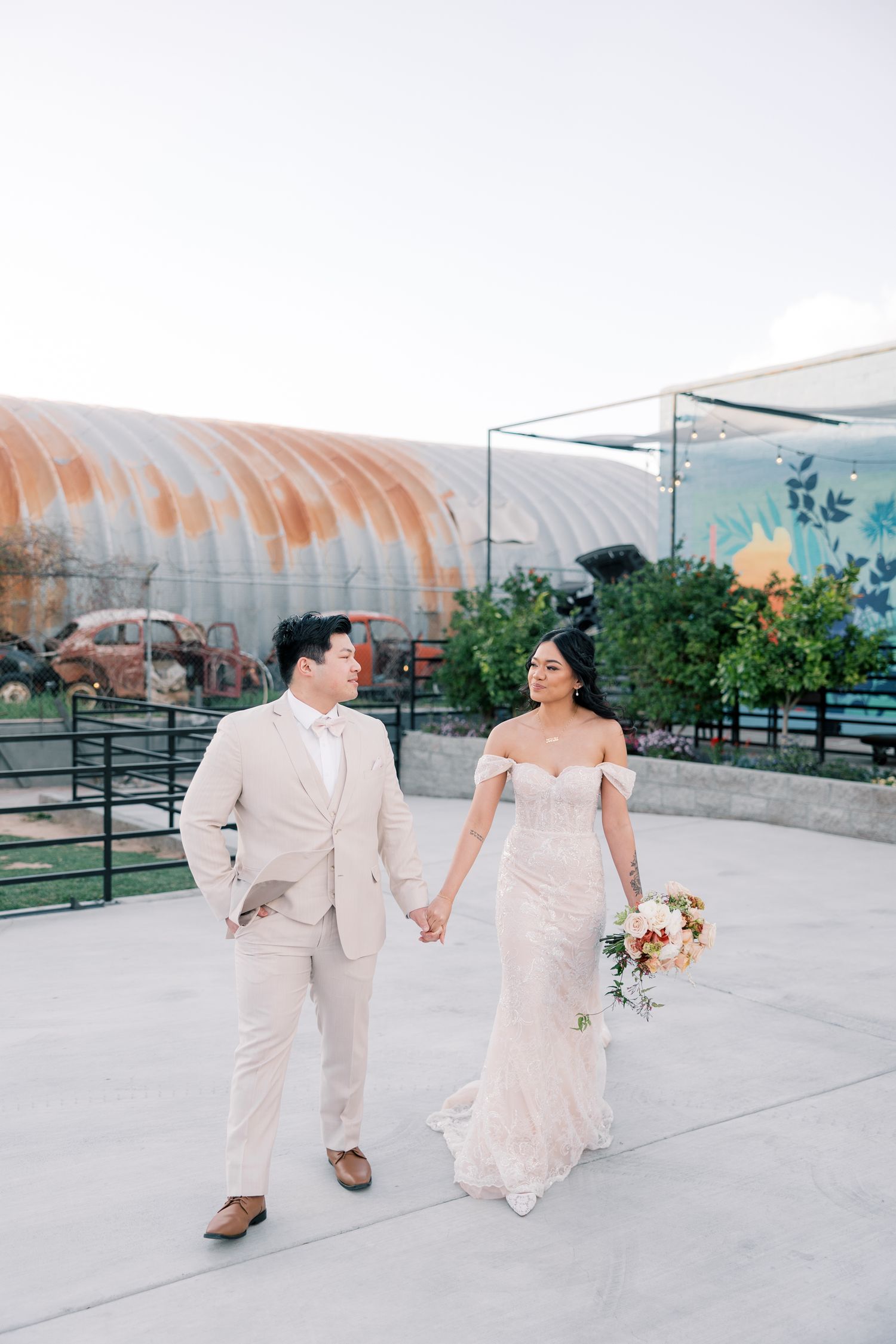 A couple in wedding attire walks together on a patio with an industrial-style building in the background.