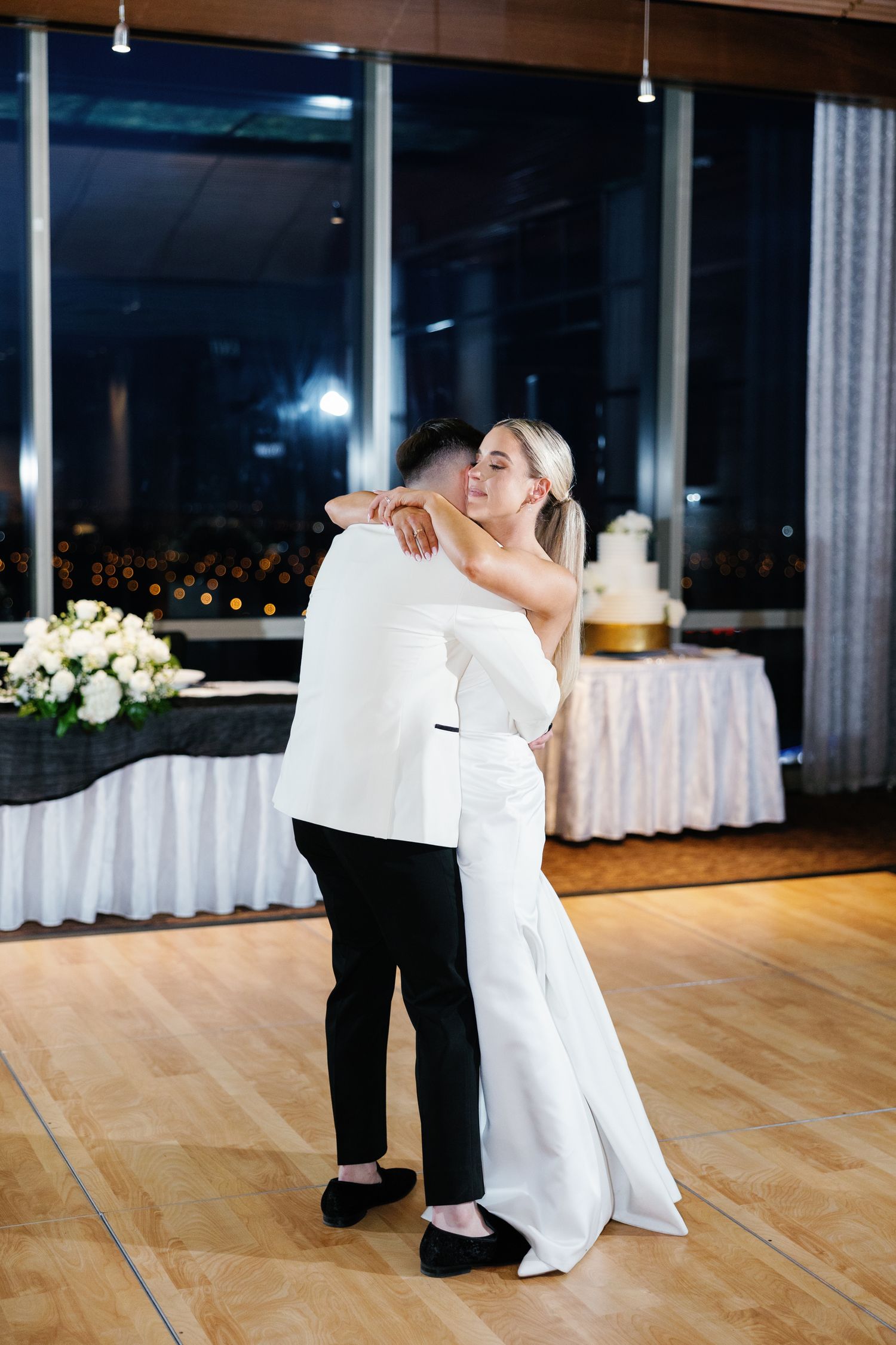 Bride and groom share first dance embrace in elegant reception hall with wedding cake in background.