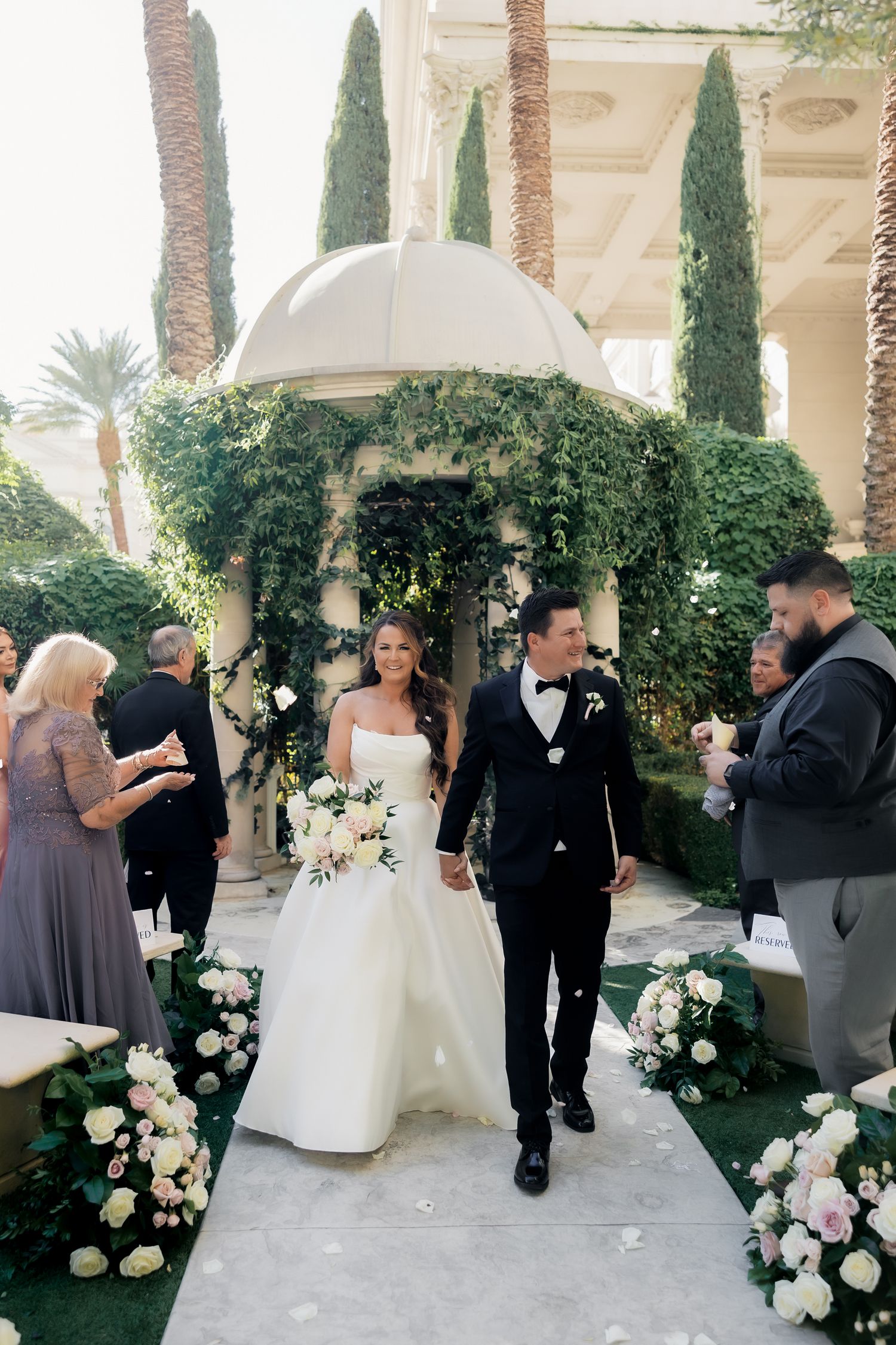 Wedding party members gather in an elegant outdoor venue with cypress trees and floral arrangements.