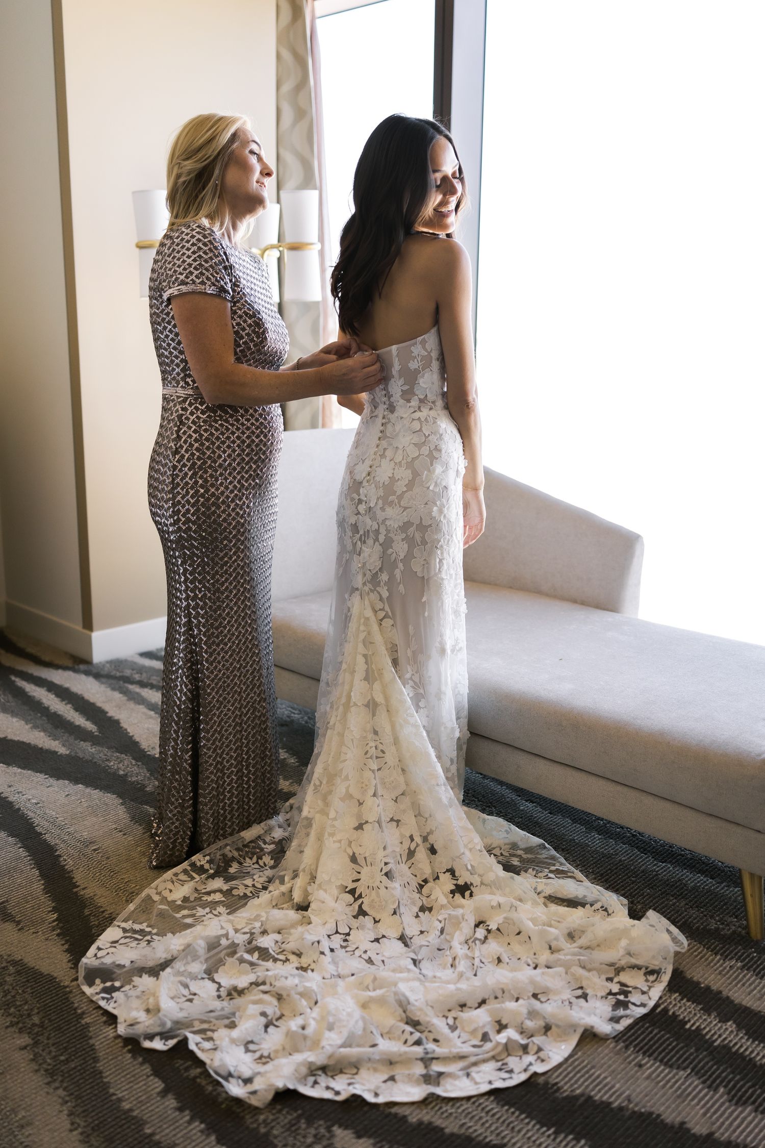 Bride in lace wedding gown being helped by woman in silver dress near window.