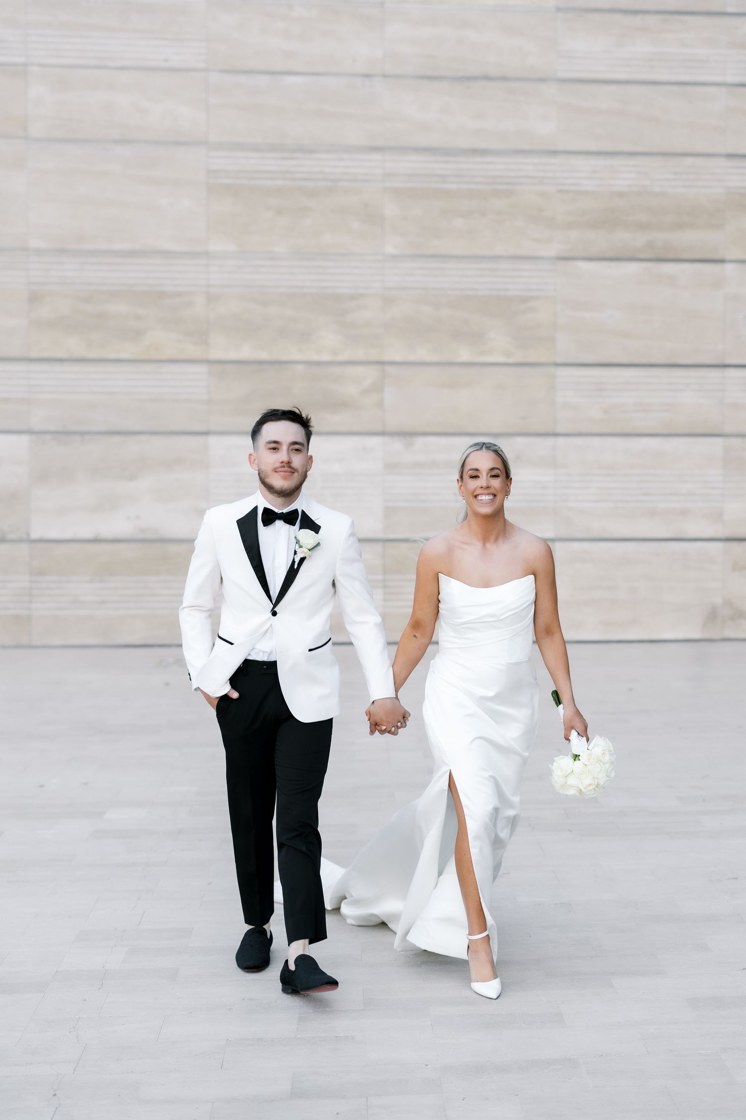 A bride and groom walk hand in hand, she in a white strapless gown, he in a white tuxedo jacket with black trousers.