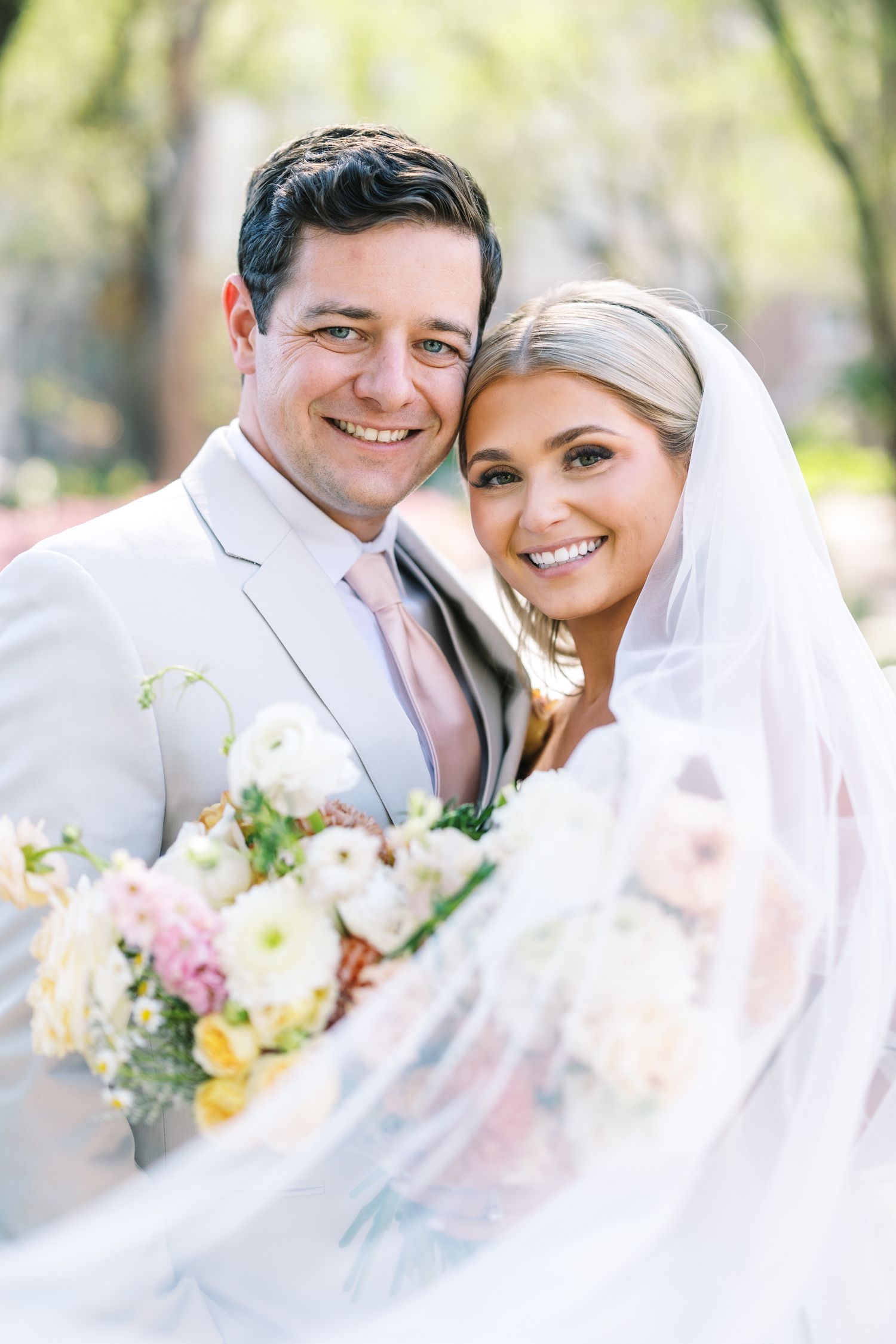 Smiling bride and groom pose together outdoors, bride holding pastel bouquet with veil flowing, groom in light gray suit.