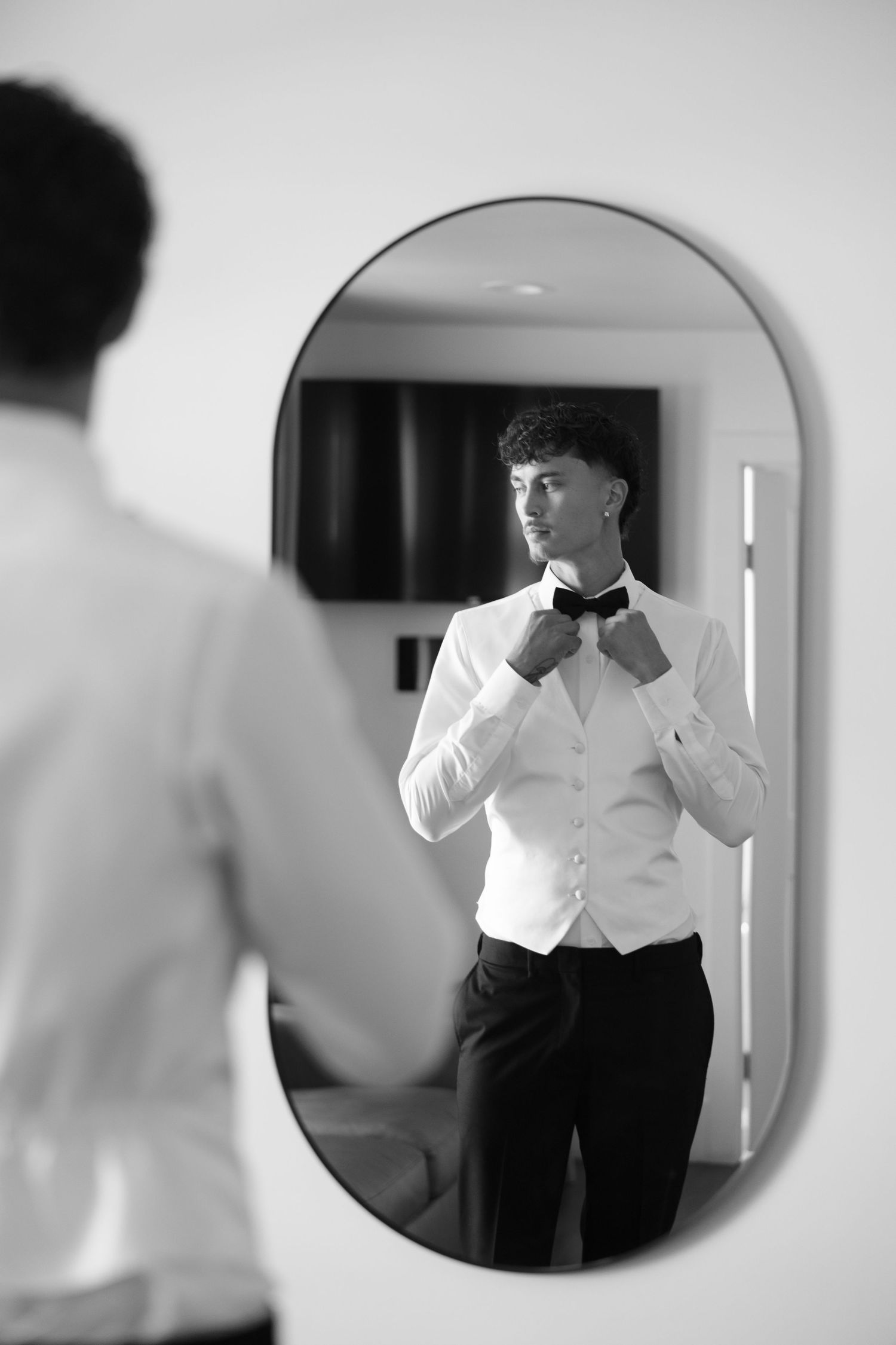 Black and white photo of a groom adjusting his bow tie while looking in an oval mirror.