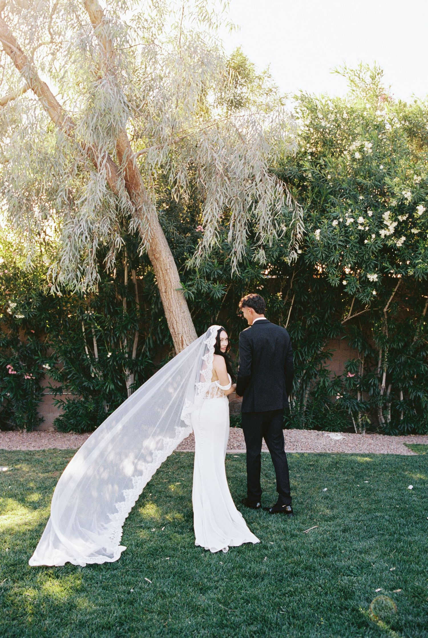 Bride in white gown with long lace veil stands with groom in black tuxedo during outdoor first look ceremony.