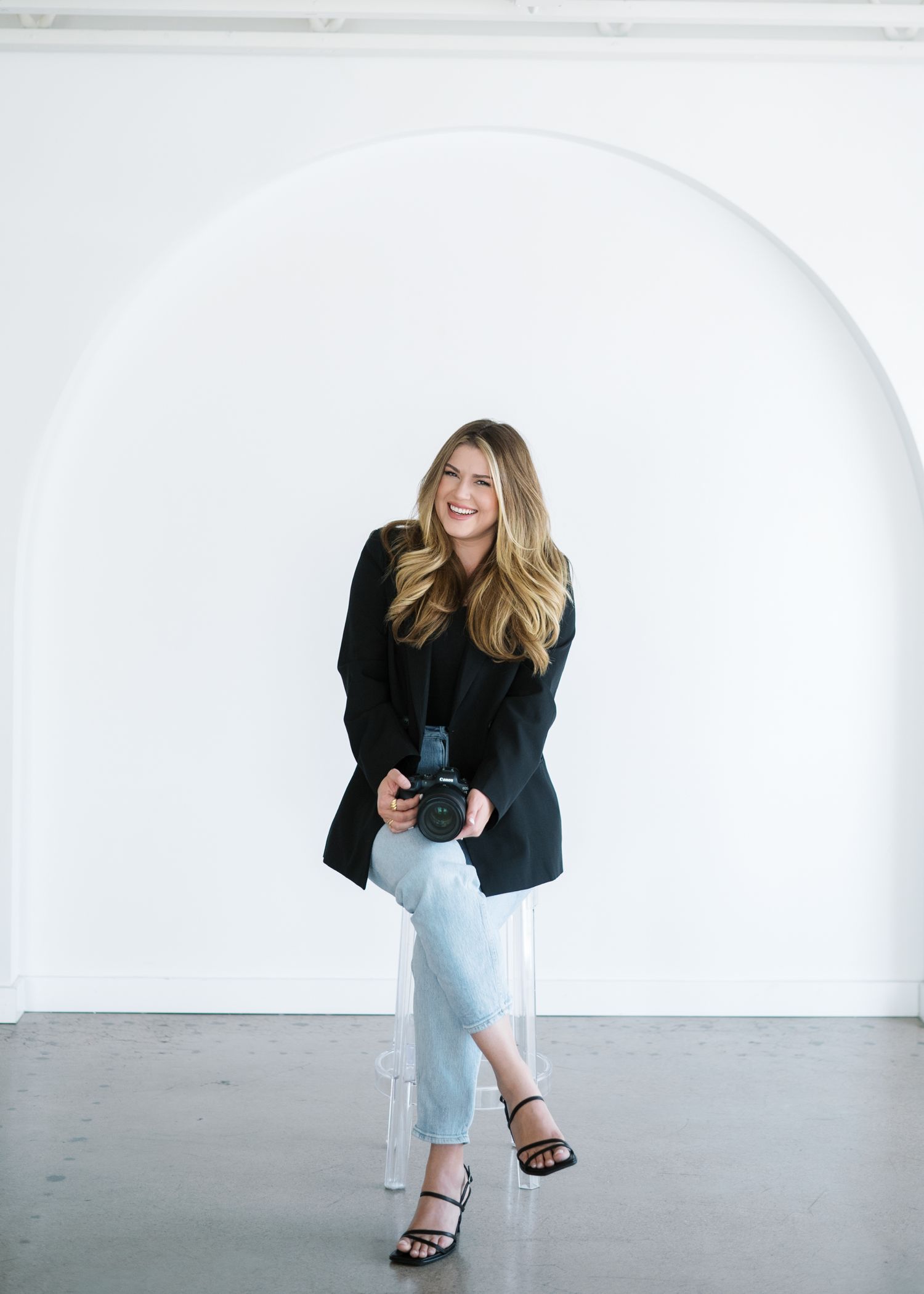 A person wearing light blue jeans and a black top poses in a white studio space with natural lighting.