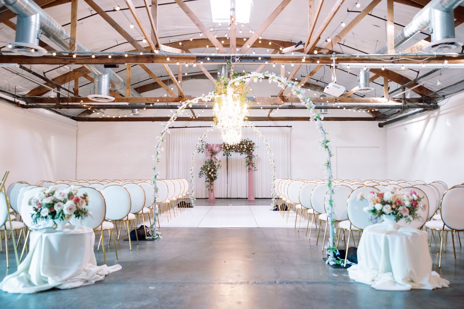 Elegant wedding venue interior with crystal chandeliers and white draped fabric decorations in an industrial-style space.