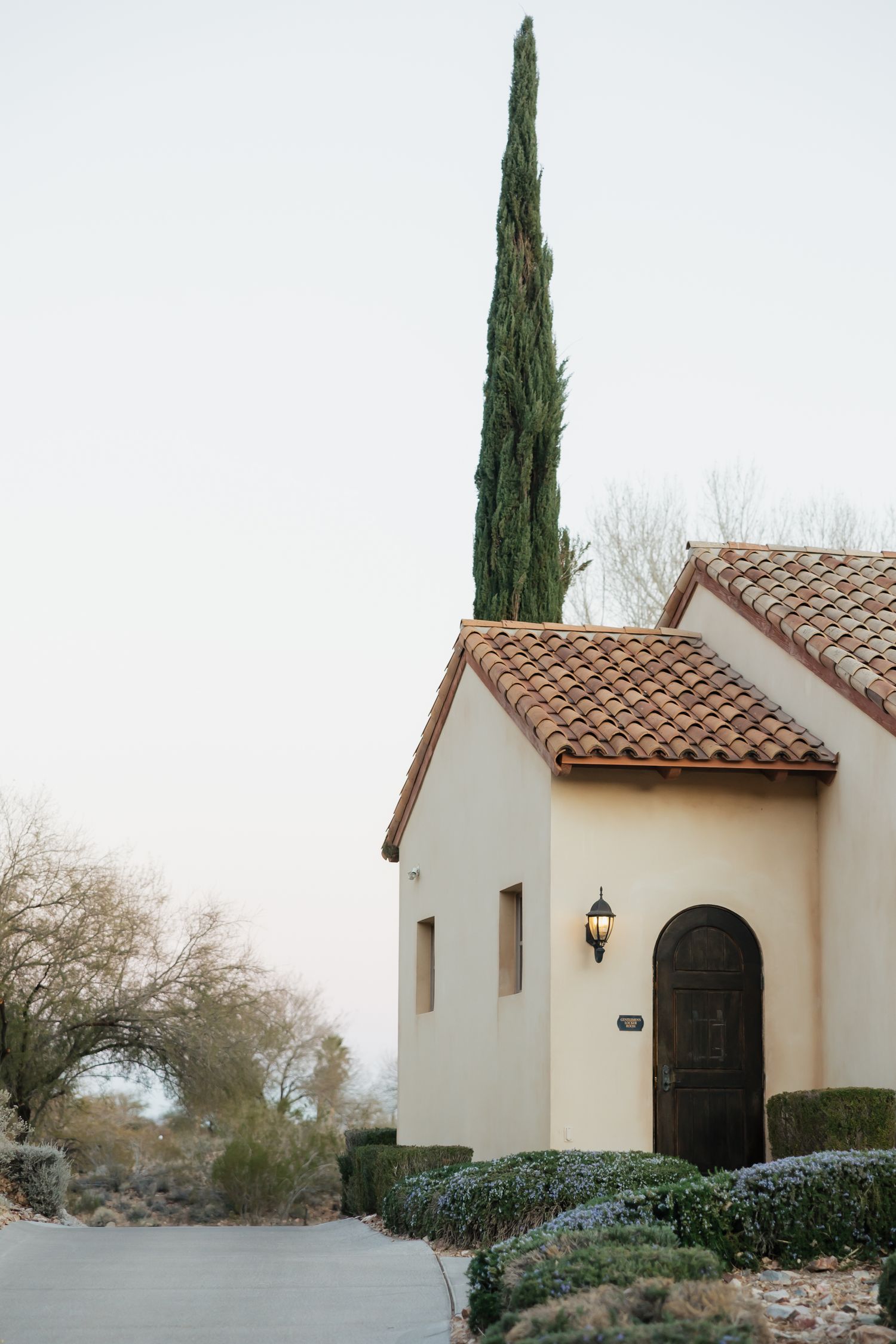 Mediterranean-style stucco home with terracotta tile roof, arched door, and tall cypress tree in a desert landscape.