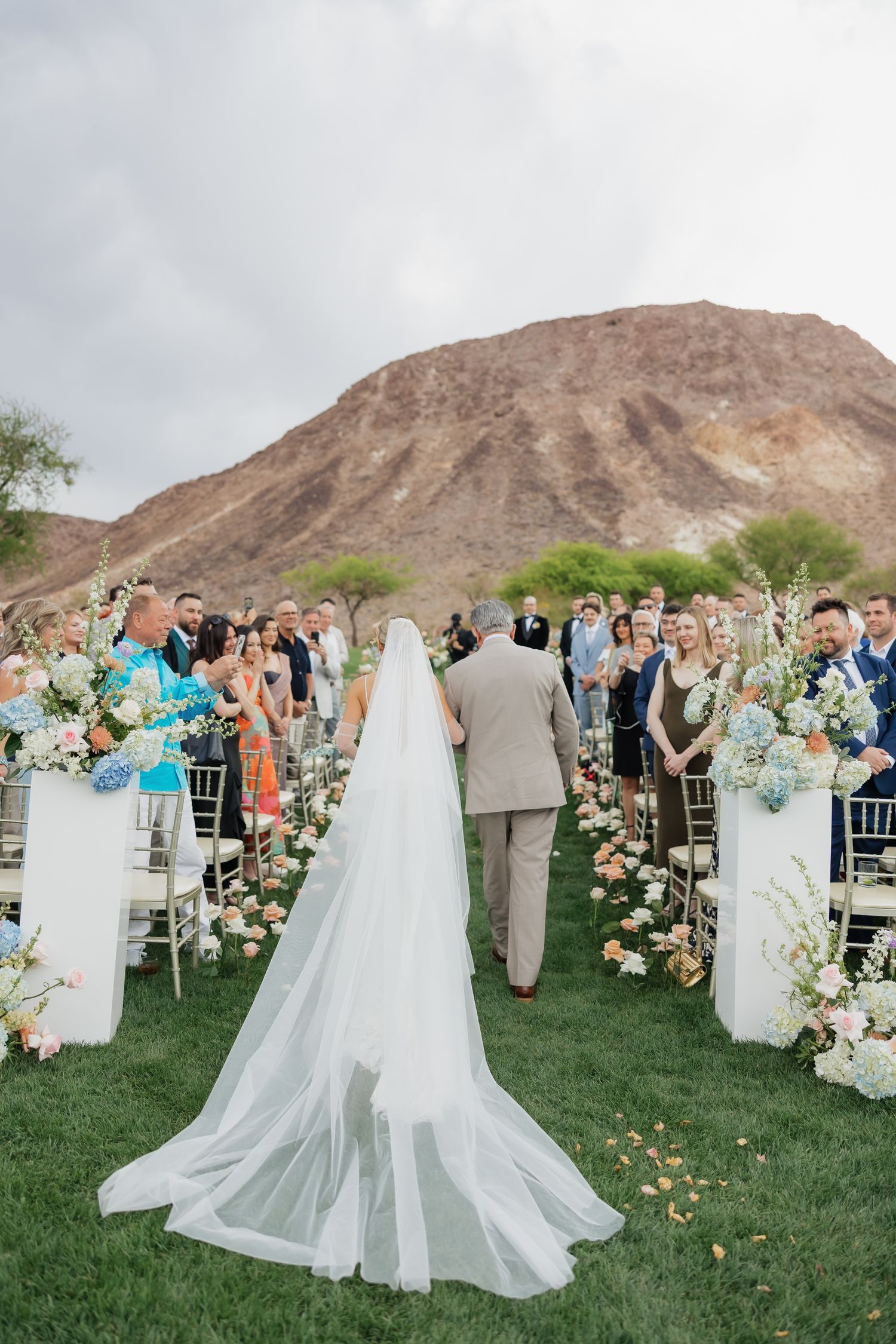 Bride walking down outdoor aisle with long veil, guests lining path, floral arrangements, desert mountain backdrop.