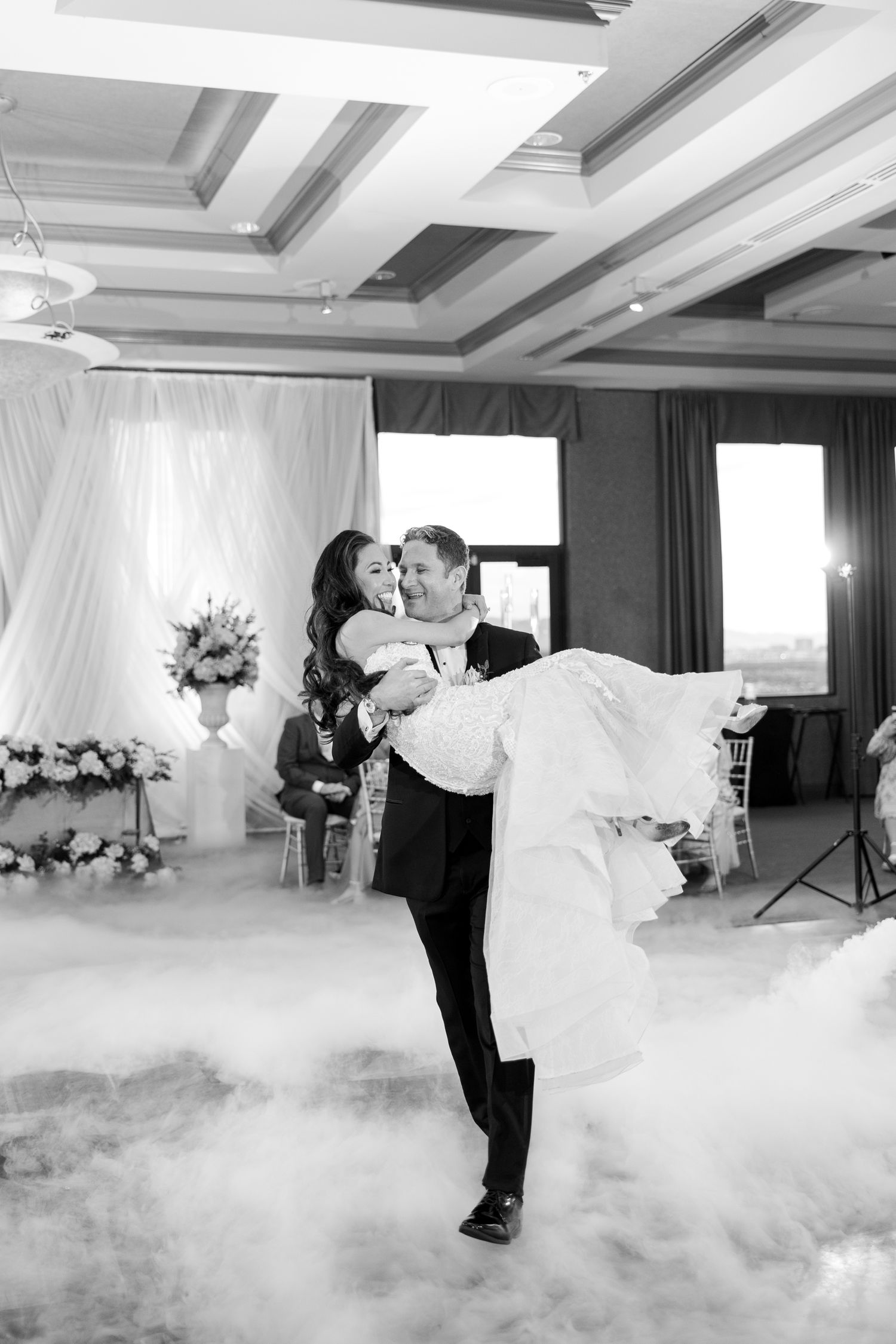 Groom lifts bride in elegant ballroom filled with fog during romantic first dance at wedding reception.