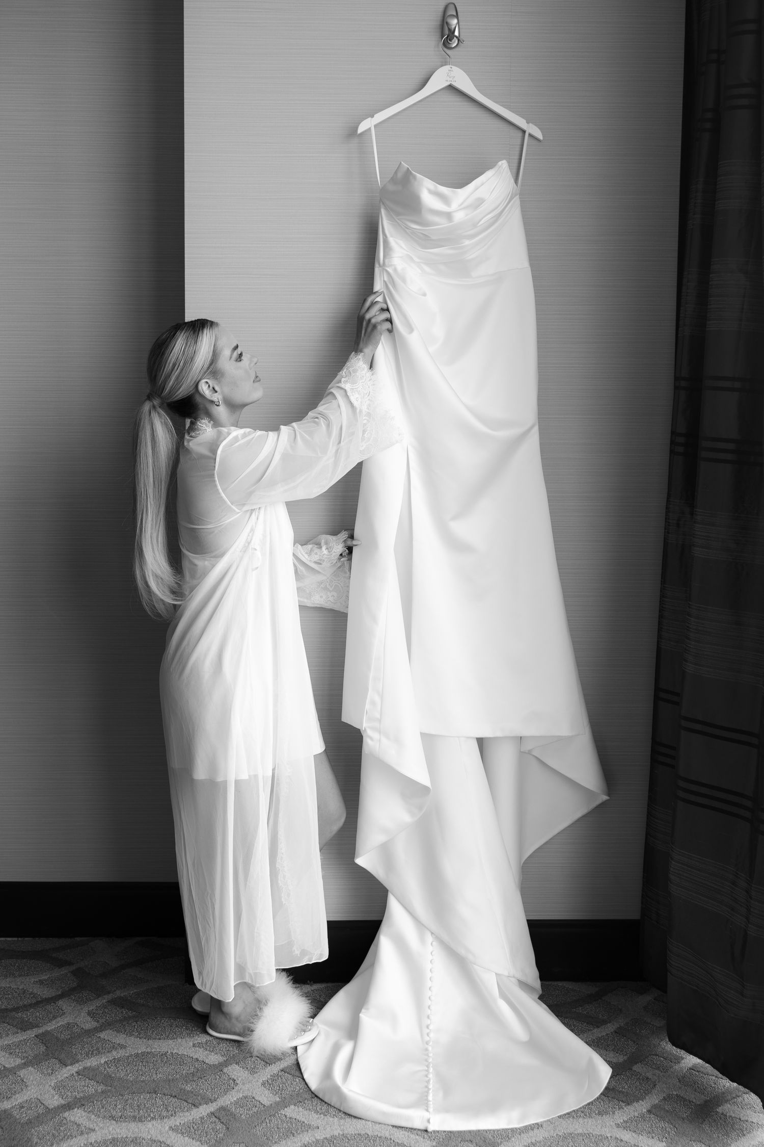 Bride in white robe admiring her elegant wedding gown hanging on a wall hook in black and white photo.