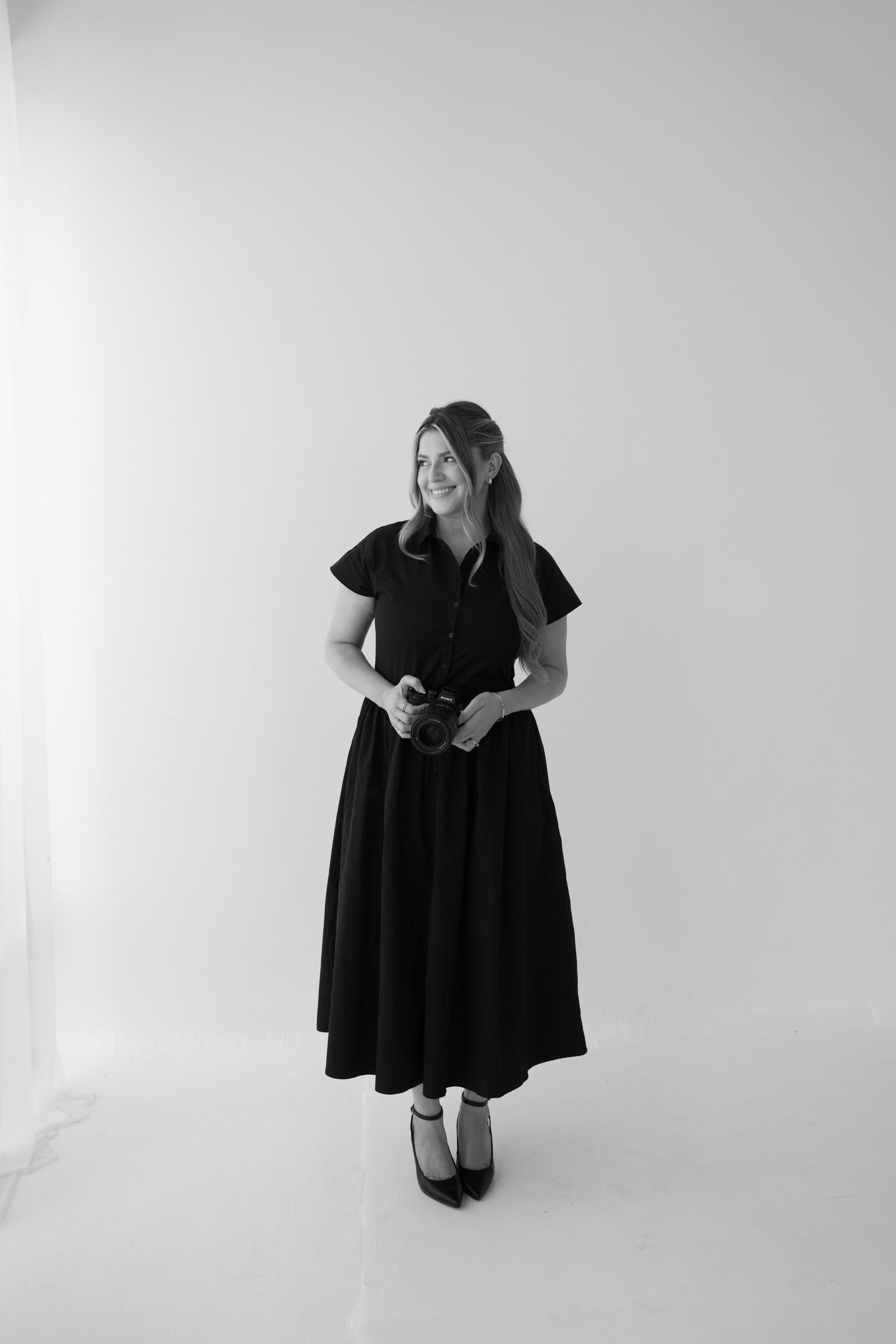 Black and white photo of a smiling woman in a black midi dress holding a camera, standing against a white background.