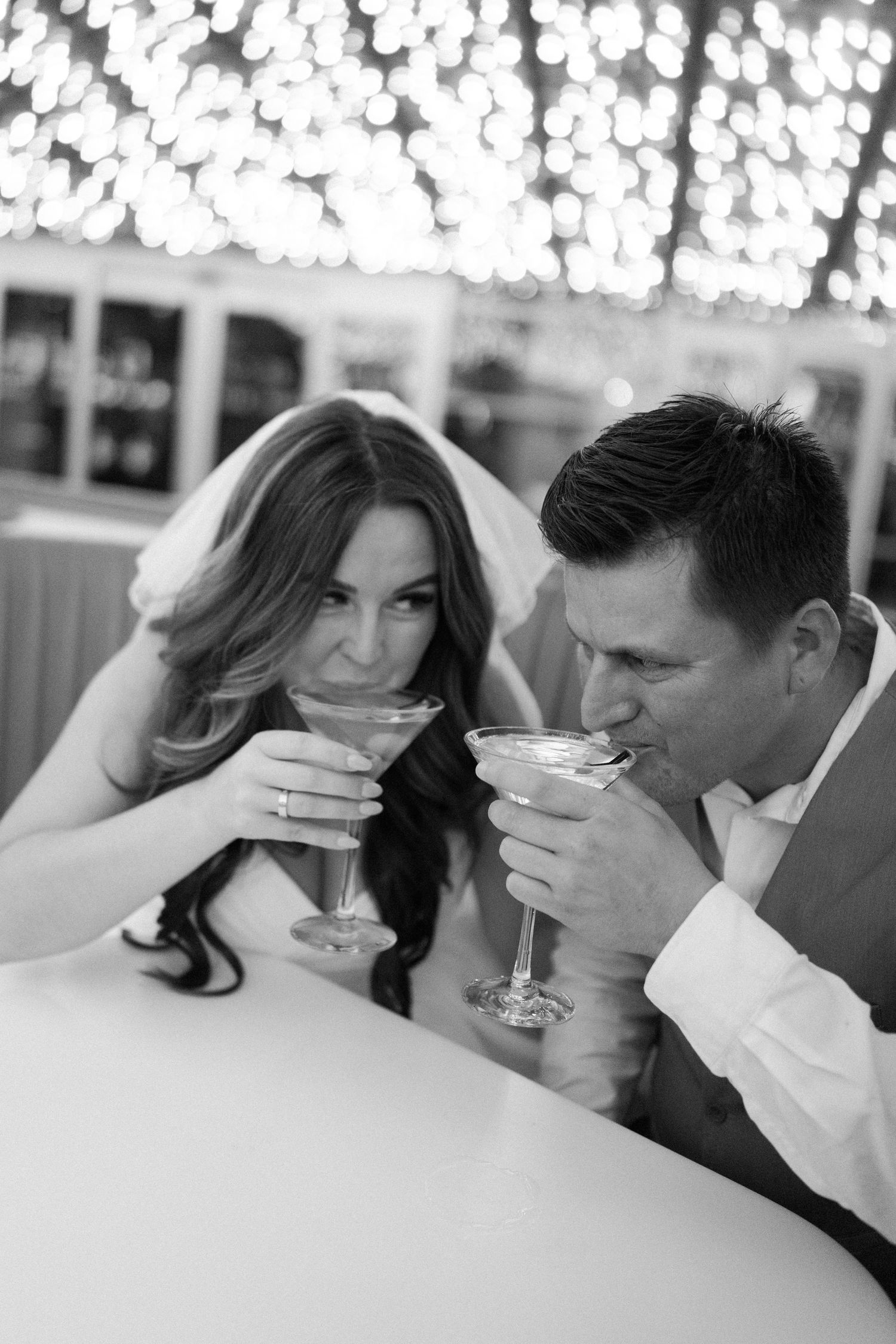 Black and white photo of a bride and groom sharing a romantic toast with martini glasses at their wedding reception.
