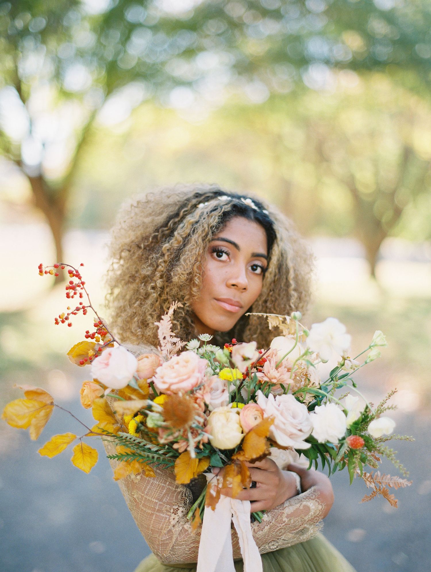 A woman with curly hair holds a lush autumn bouquet of white, peach, and yellow flowers outdoors.