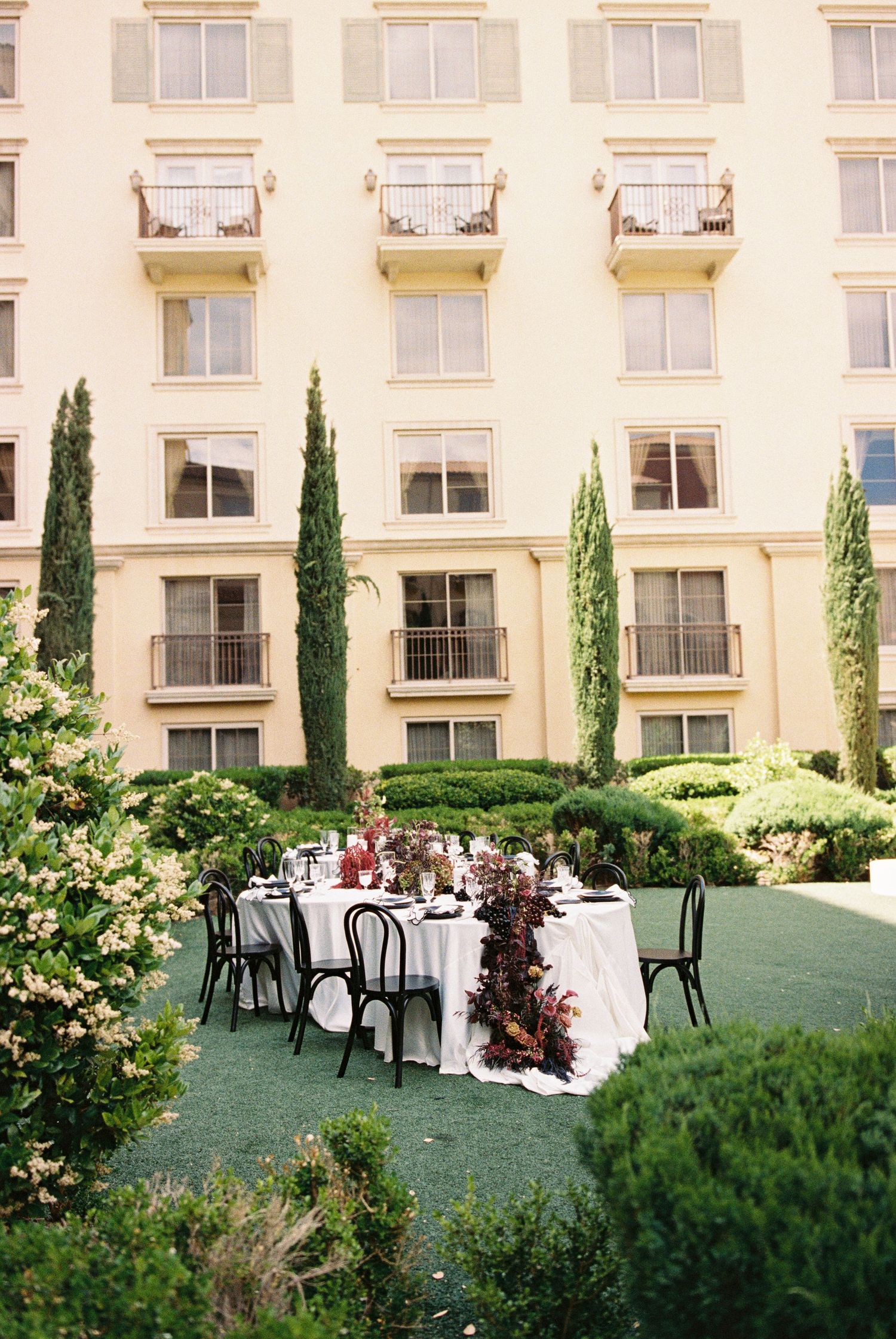 Elegant outdoor wedding reception table set in a hotel courtyard garden with lush greenery, dark florals, and black bistro chairs.