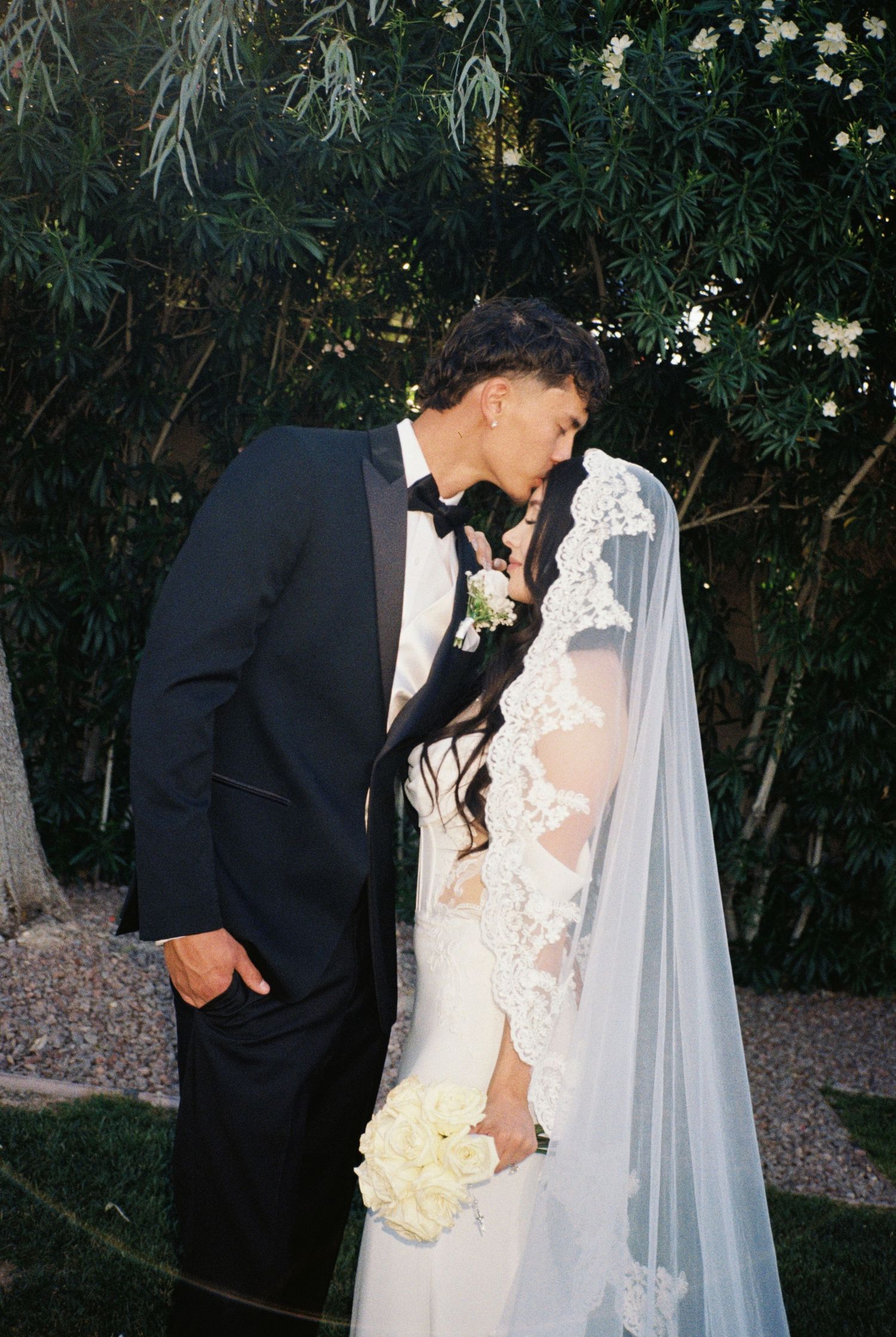 A bride and groom share a romantic kiss outdoors, she in a lace wedding gown with veil, he in a black tuxedo.