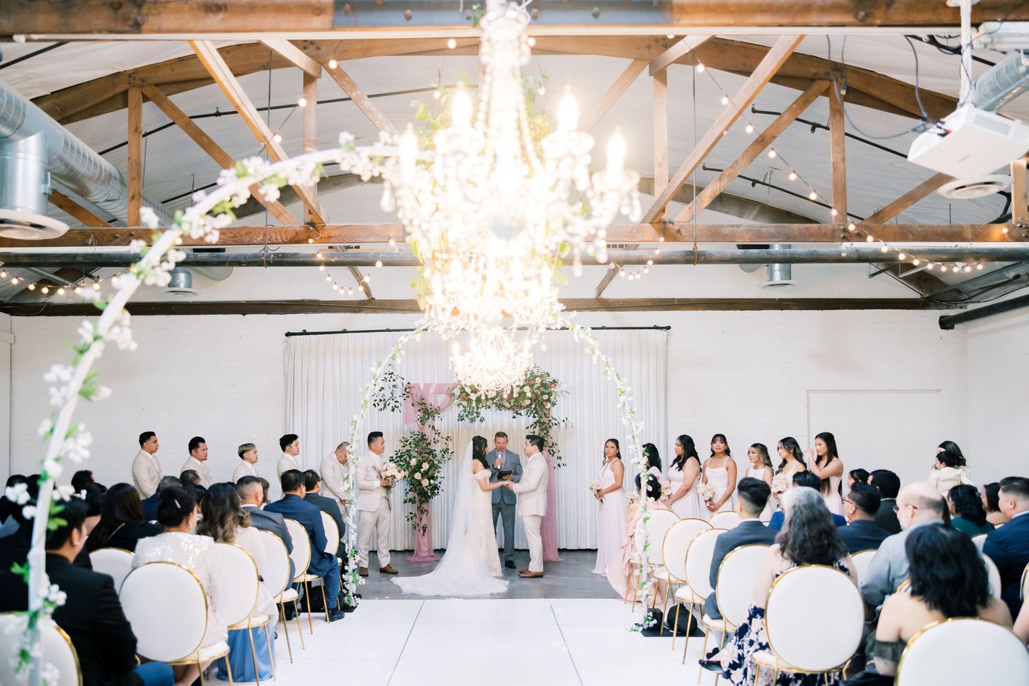Wedding ceremony setup in a bright white industrial space with chandeliers and rows of guest seating.