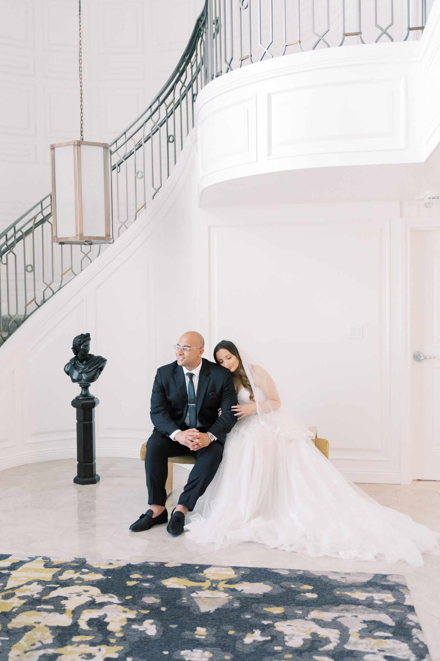 Bride and groom sitting together in elegant white mansion foyer with curved staircase and ornate decor.