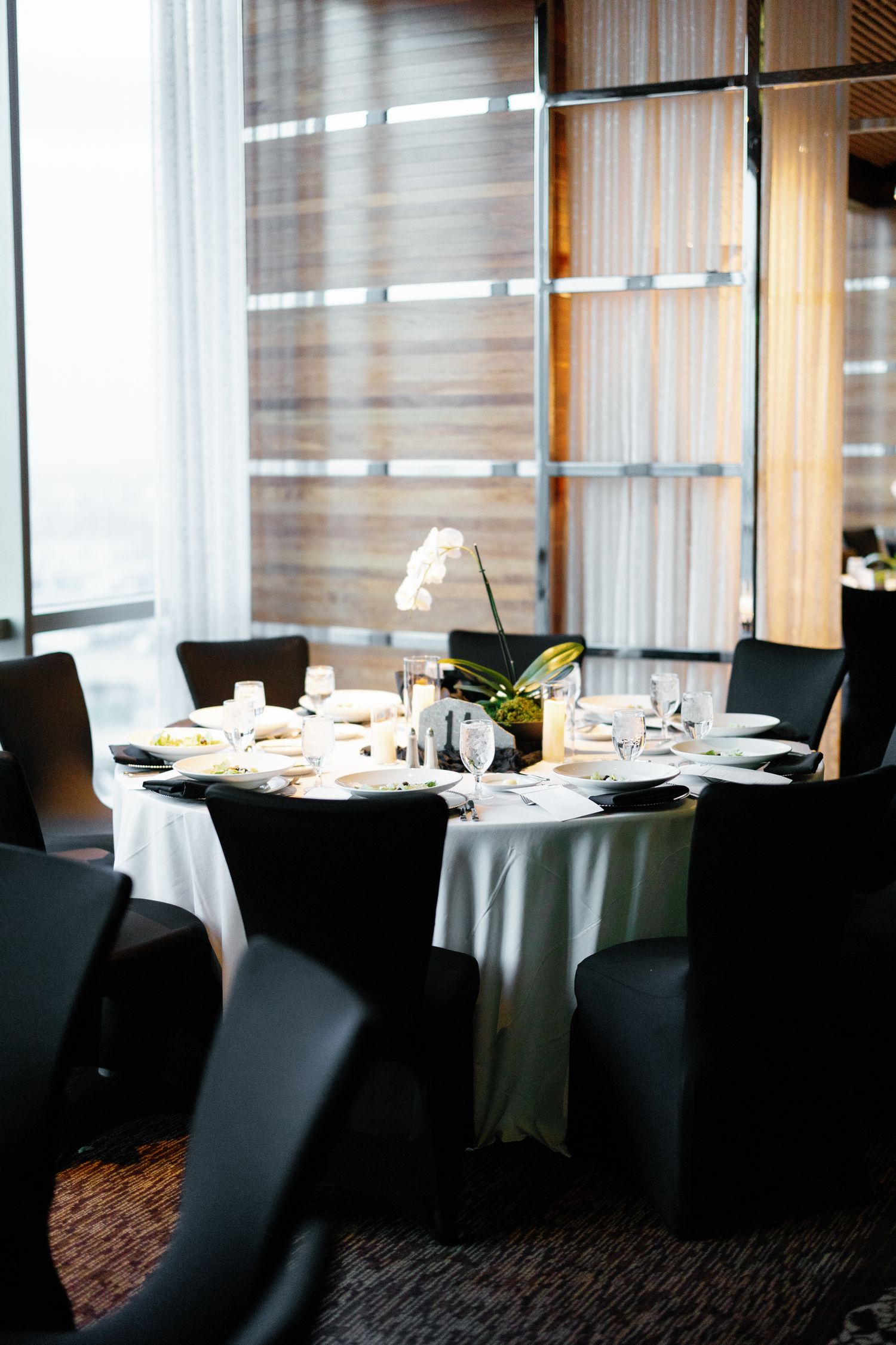 Elegant round banquet table with white linen, black chairs, glassware, and white orchid centerpiece in a modern venue.