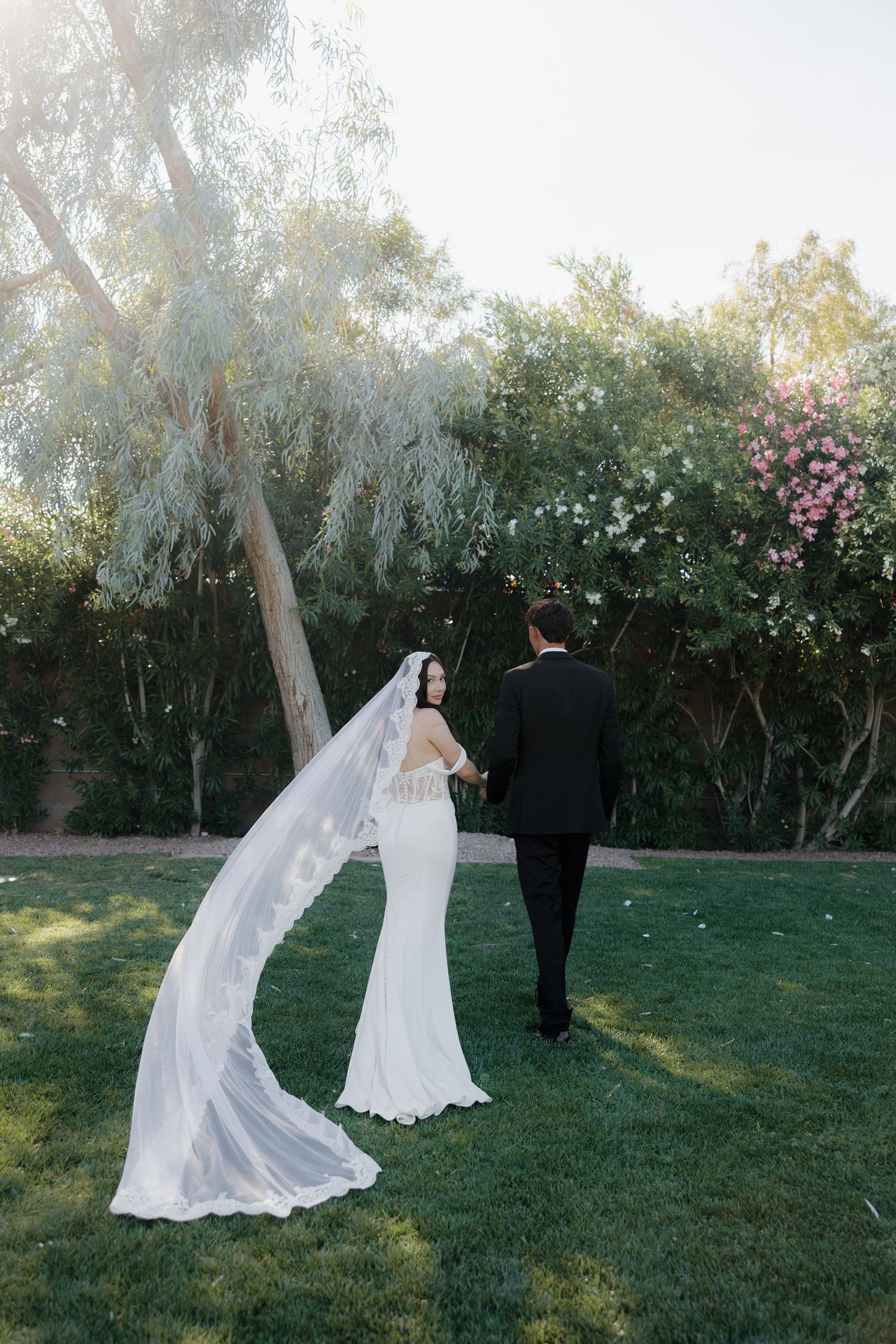 Bride in white gown with long veil holds grooms hand walking through lush garden on wedding day.