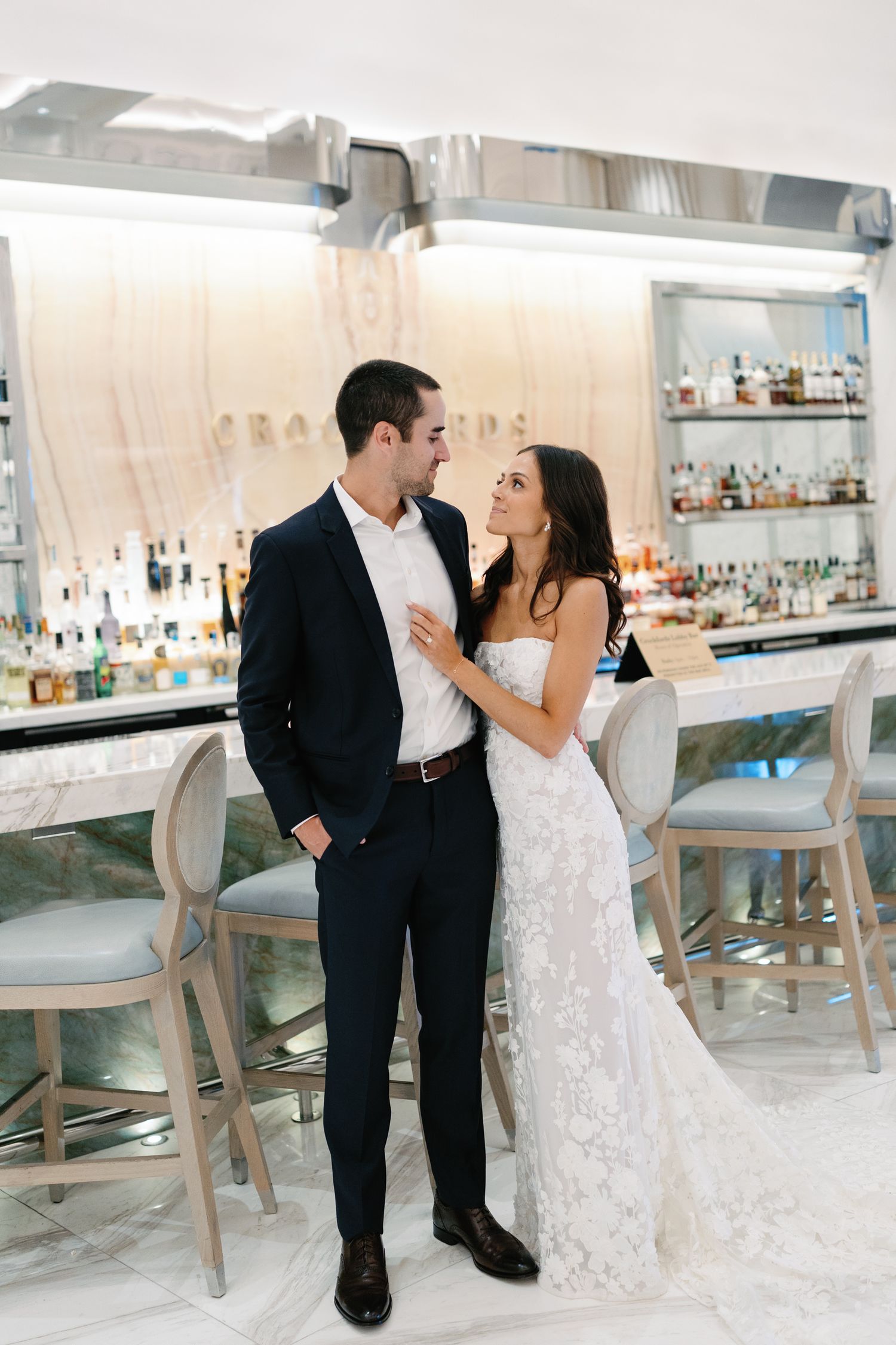 Bride in lace gown and groom in navy suit pose together at an elegant marble bar during their wedding reception.